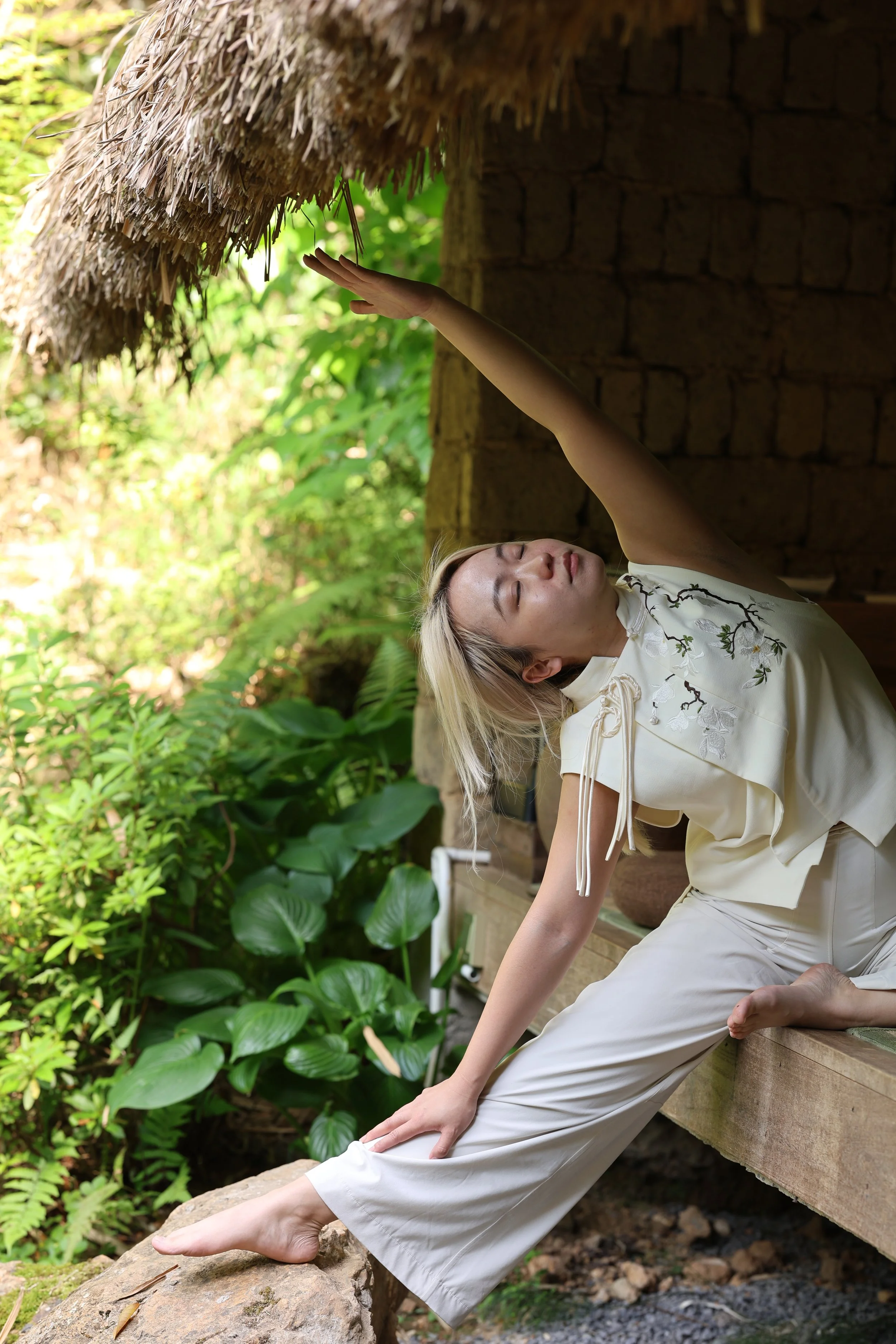Evelyn in a side stretch beneath a thatched roof, surrounded by greenery