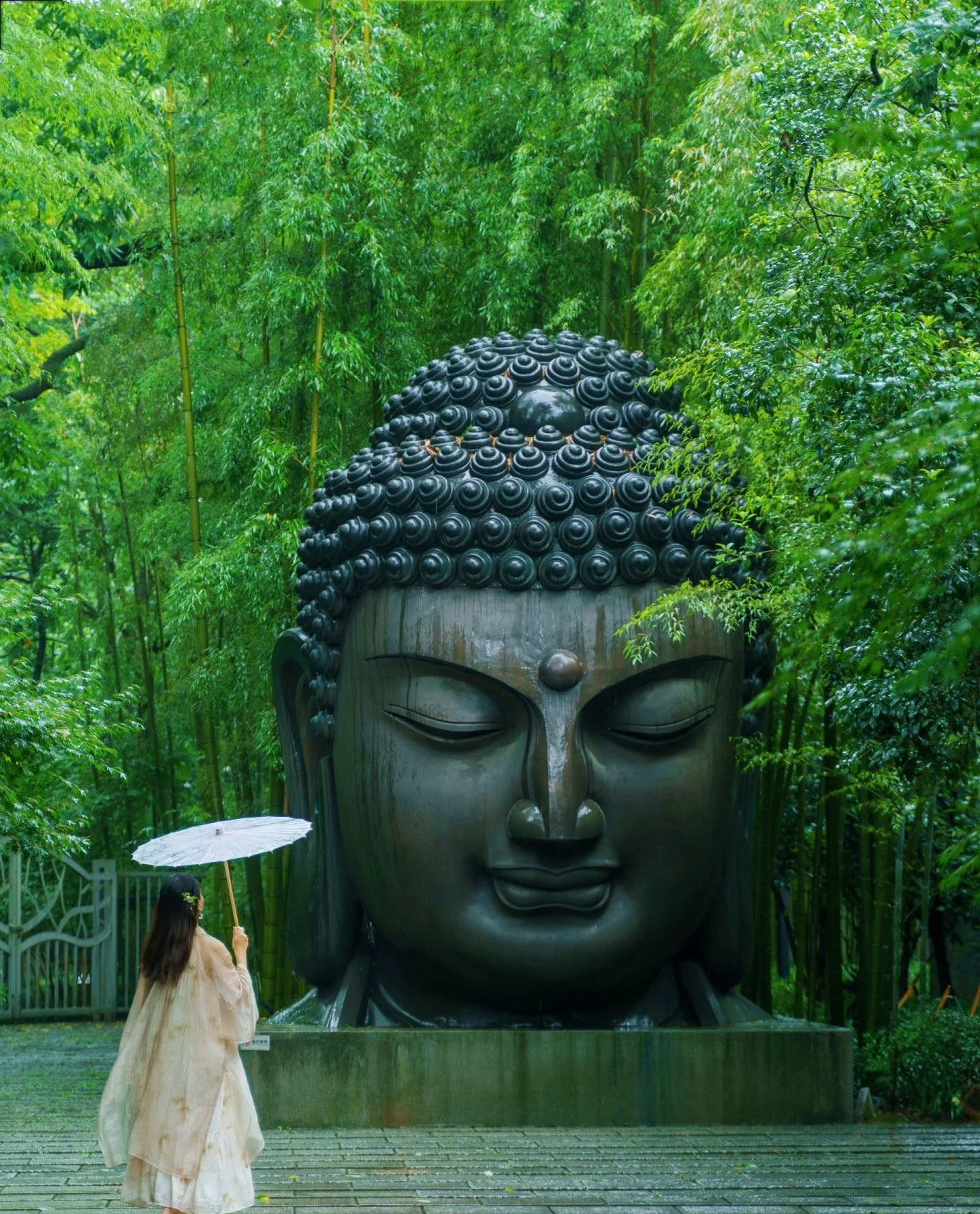 A large Buddha head sculpture surrounded by lush green trees with a person holding a white umbrella standing in front of it.
