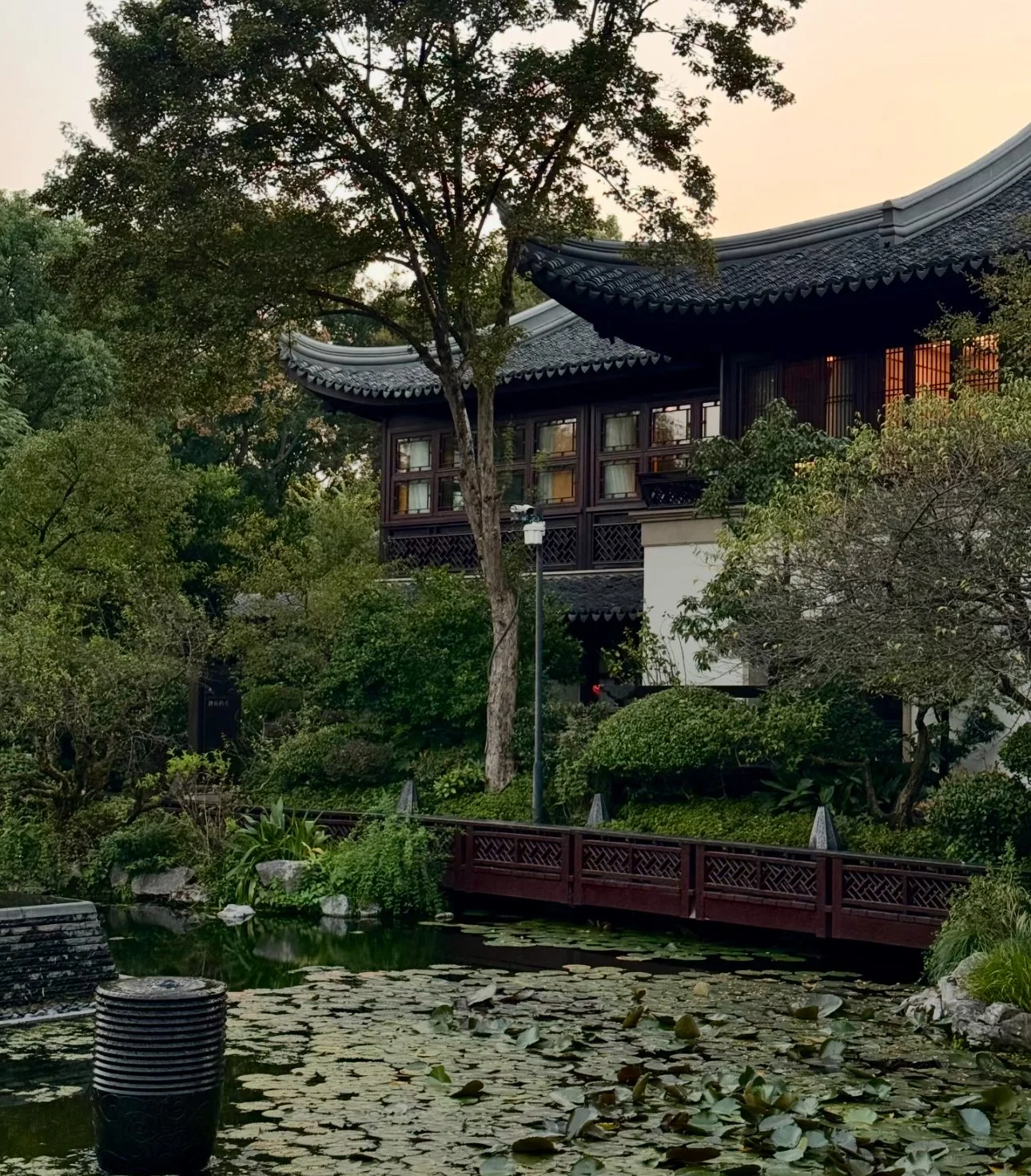 Traditional Asian-style building with curved roof, surrounded by lush greenery and trees, beside a pond with lily pads and a wooden bridge.