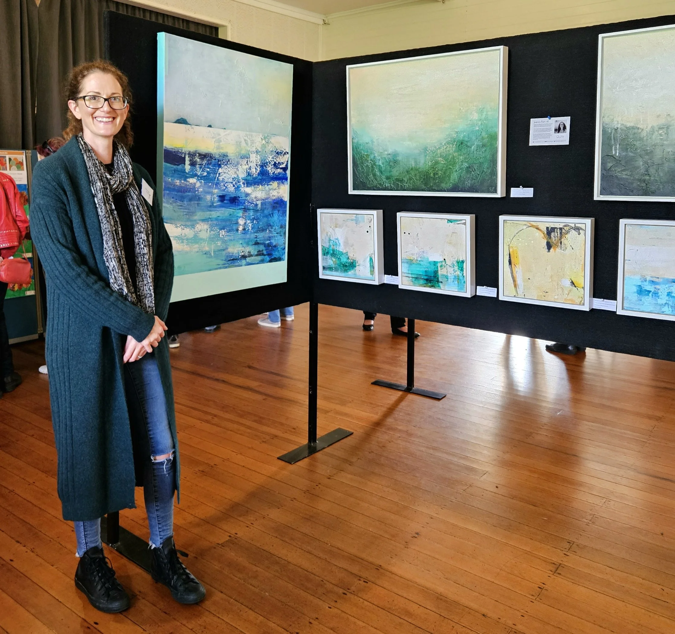 A woman with glasses and curly hair standing in front of a black display wall at an art gallery, smiling. The wall behind her shows several abstract paintings with various shades of blue, green, and white. The gallery has wooden floors and other visitors are visible in the background.