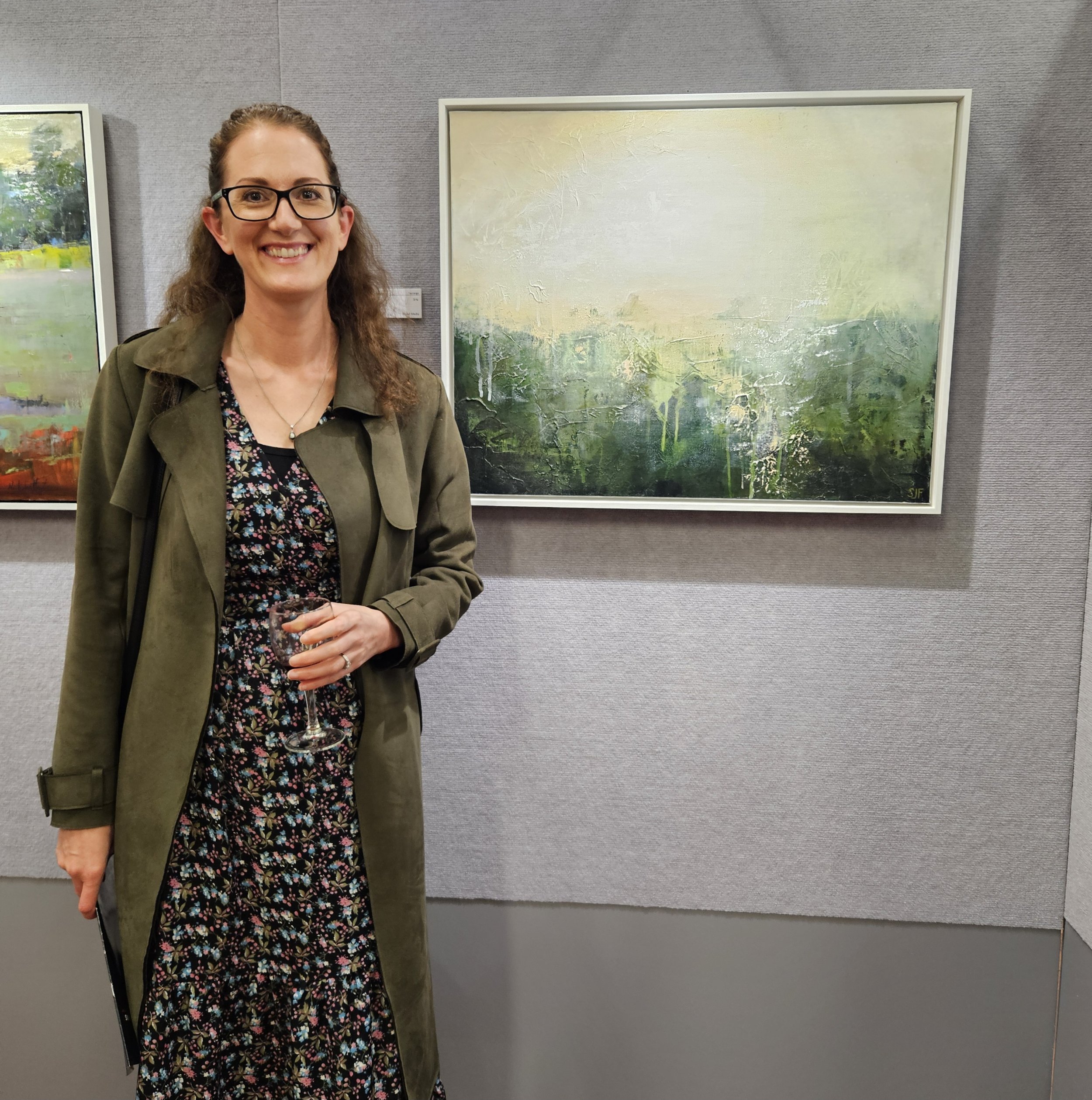 Woman with glasses and brown hair smiling at an art gallery, standing in front of an abstract landscape painting with green, yellow, and blue hues.