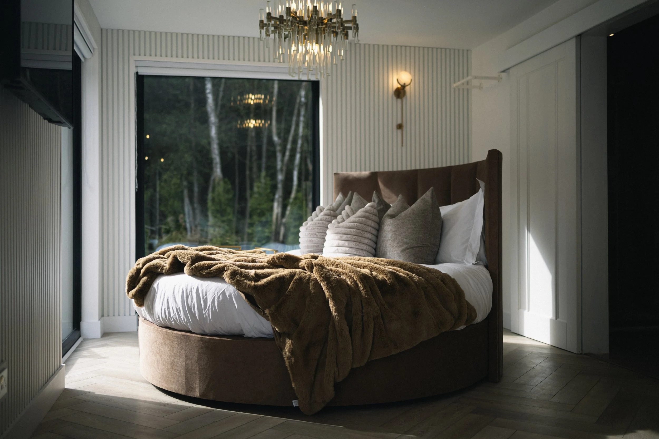 A cozy bedroom with a large round bed covered in white sheets, adorned with multiple pillows and a plush fur blanket. The room features a large window showing a forest outside, a modern chandelier hanging from the ceiling, and a wall-mounted light fi