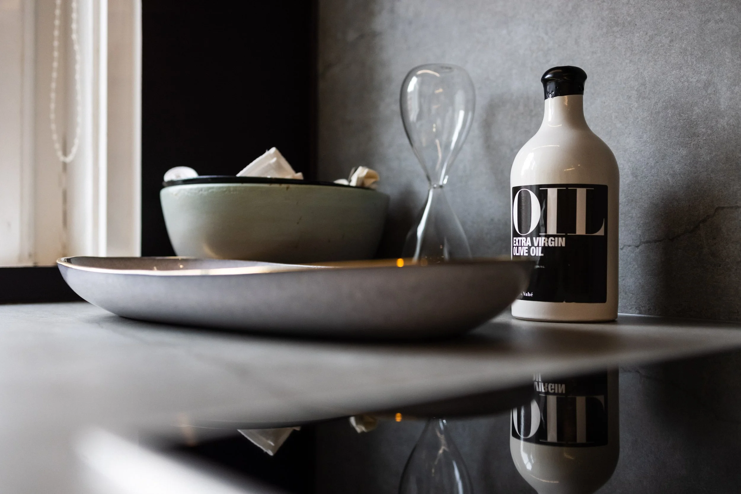 Countertop with a beige bottle of extra virgin olive oil, a glass vase, a green bowl with paper napkins, and a gray tray, against a gray wall and nearby window.