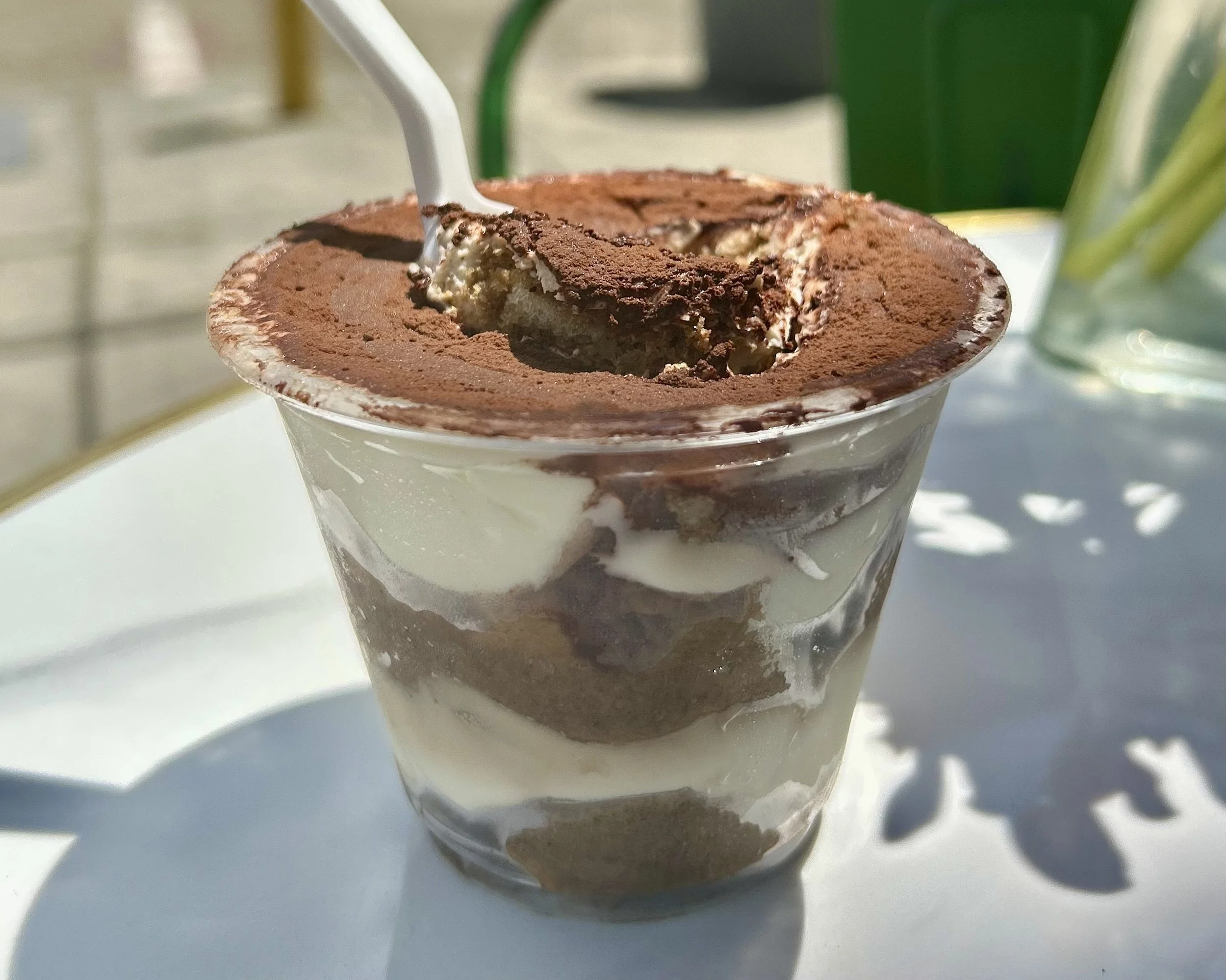 A glass cup of tiramisu topped with cocoa powder with a straw inserted.