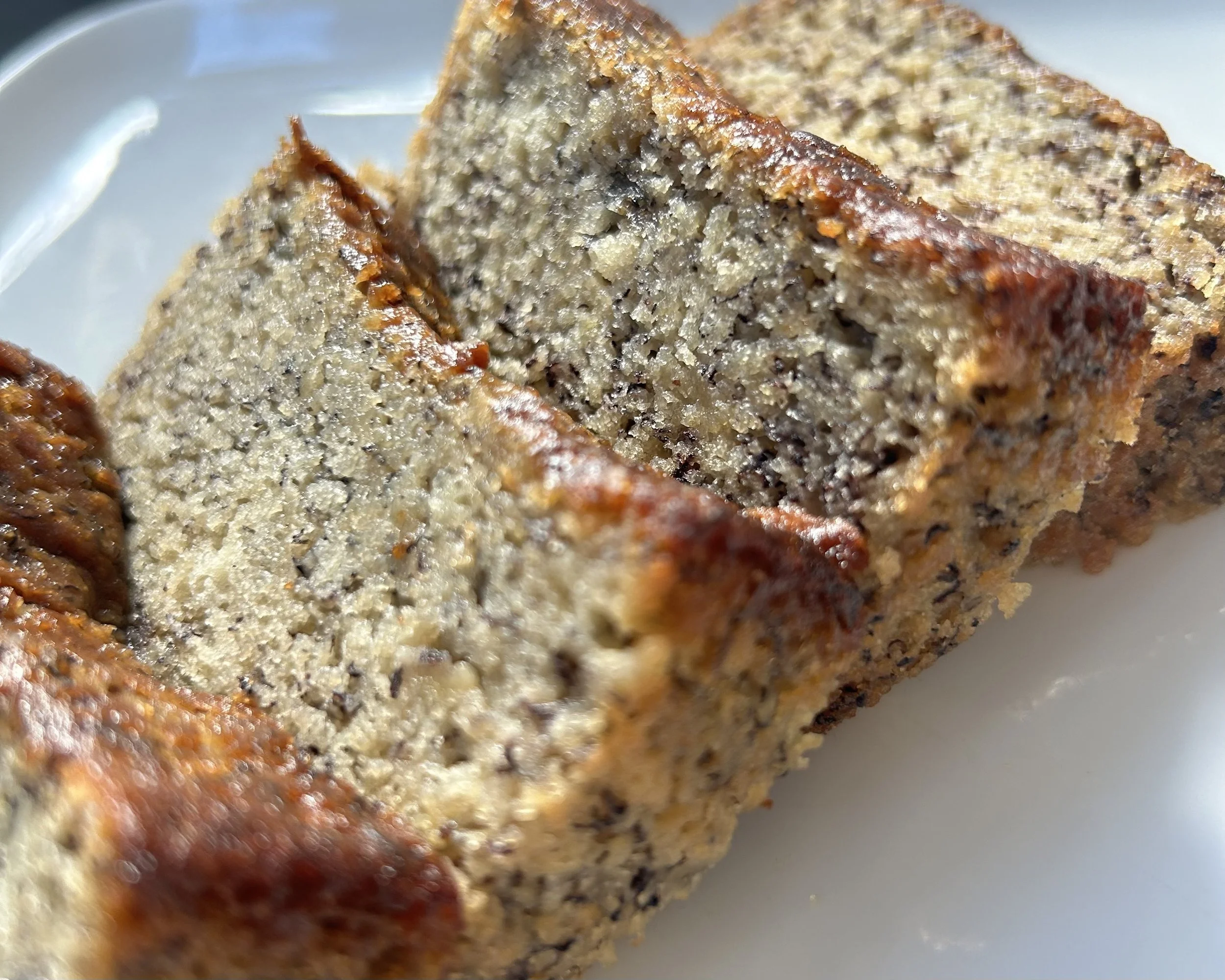 Close-up of a sliced homemade banana bread loaf with a golden-brown crust, showing its moist, dense interior with small dark specks, on a white plate.