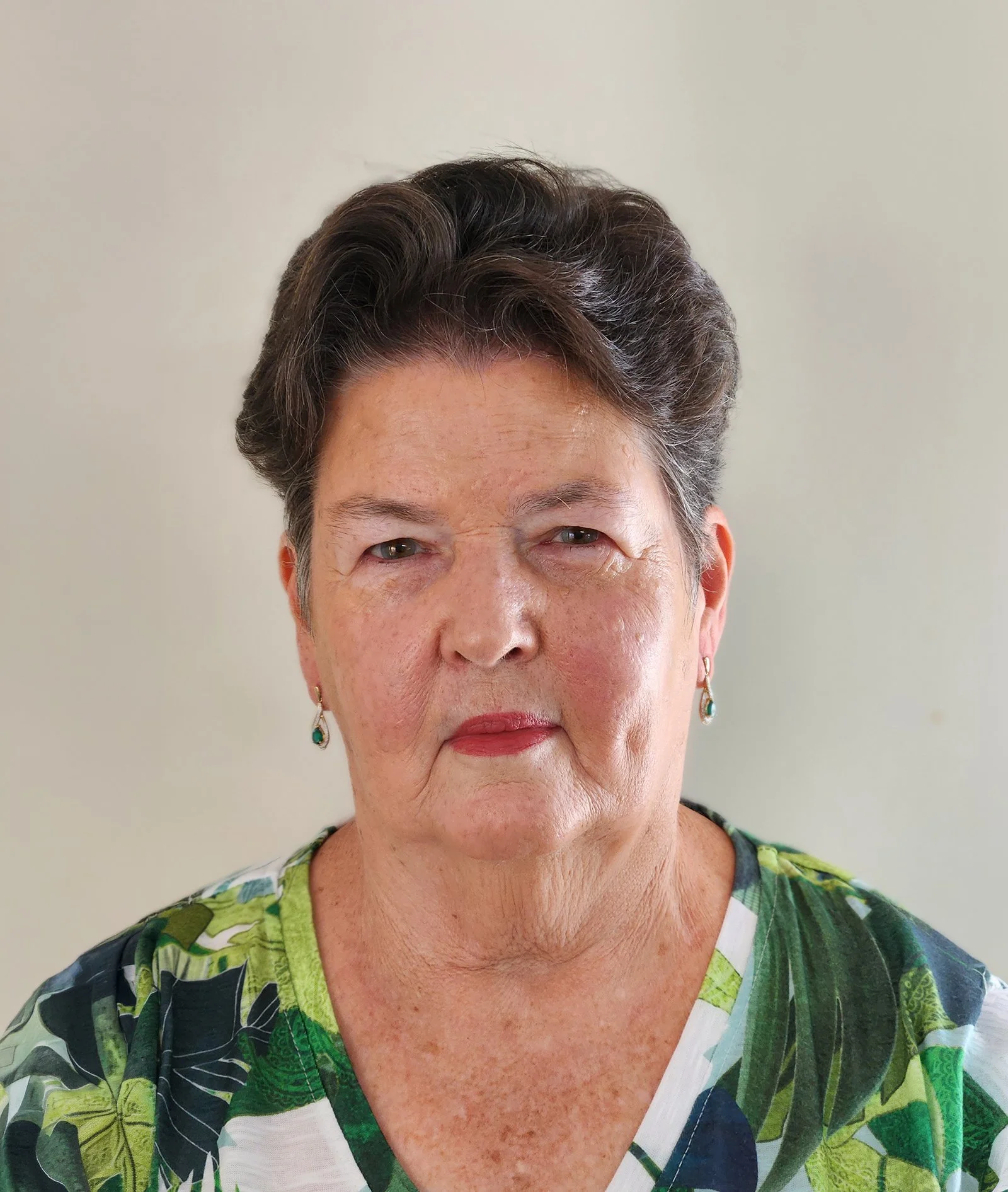 A portrait of an older woman with short, dark hair, light skin, and wearing earrings with turquoise stones, a floral blouse, and a neutral background.