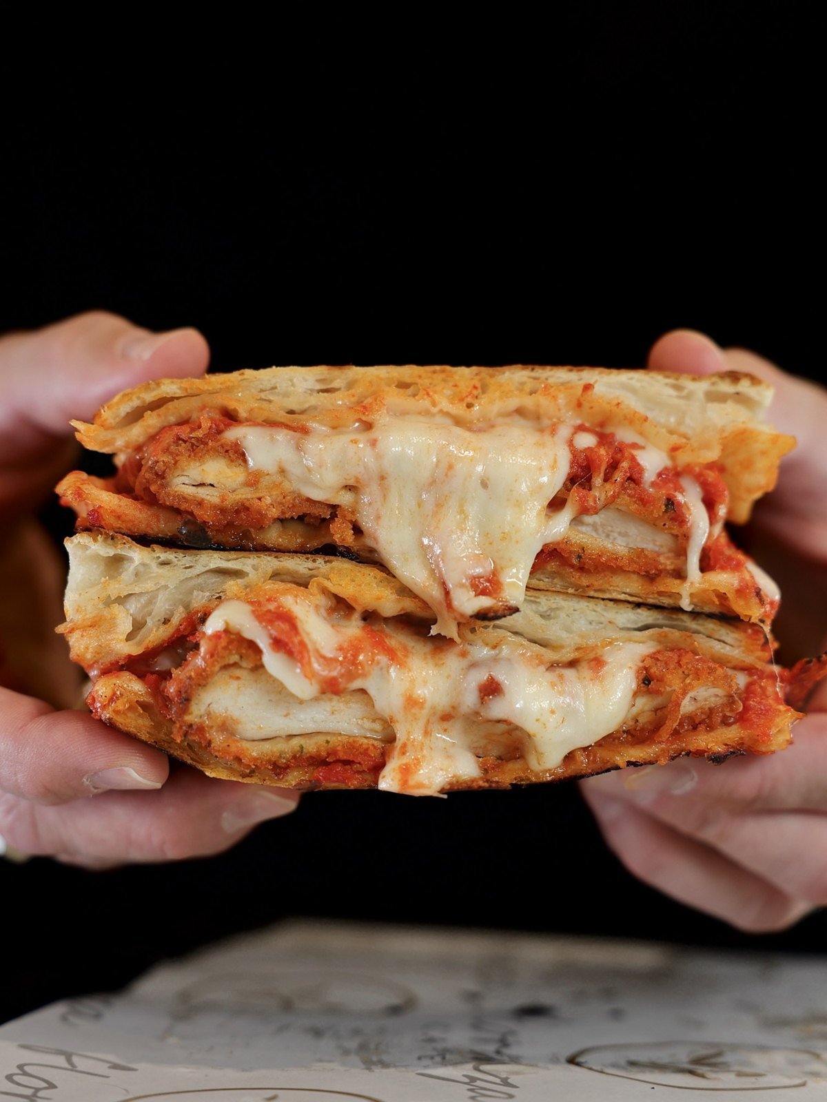Hands holding stacked chicken Parmigiana panini with tomato sauce and melted mozzarella.