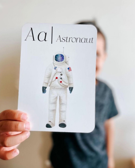Child holding a card with an illustration of an astronaut and the letter 'A' and the word 'Astronaut' in front of their face.