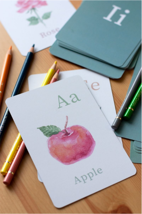 Educational flashcards for learning the alphabet, featuring a pink apple illustration and the words 'Aa' and 'Apple', with colored pens and additional flashcards visible on a wooden surface.
