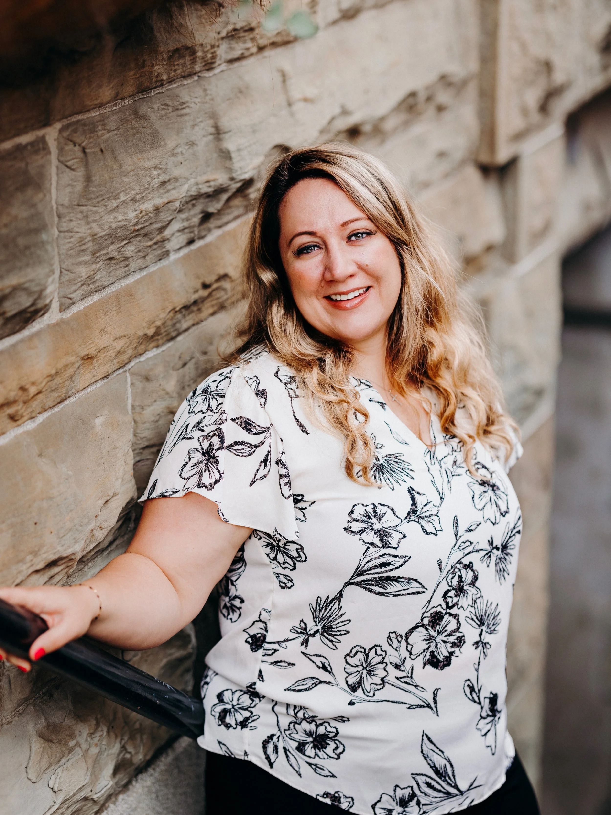 A woman with long wavy blonde hair and light skin, smiling, leaning against a stone wall. She is wearing a white blouse with black floral patterns, and holding a black object in her left hand, which is slightly visible in the lower left corner of the image.