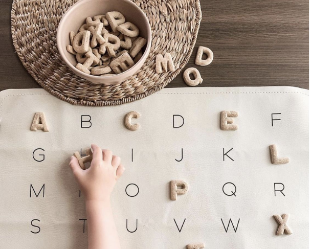 A child's hand reaching to place the letter 'H' on a chart with the alphabet written in black lines, with a bowl of letter-shaped cookies and some cookies scattered on a wicker tray.
