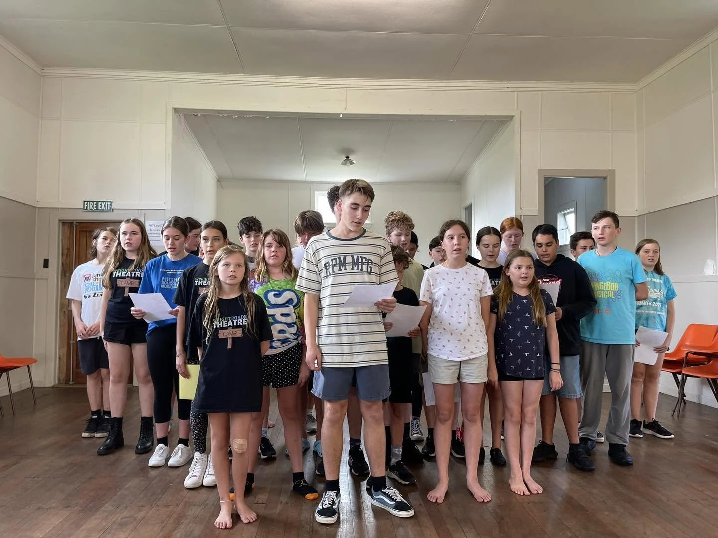 A group of children and teenagers standing in a room, with some holding papers and seemingly singing or reciting.