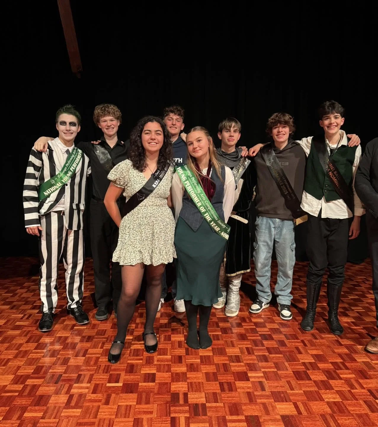 Group of seven young people at an award ceremony, wearing sashes and costumes, standing on a wooden stage with black backdrop, smiling.