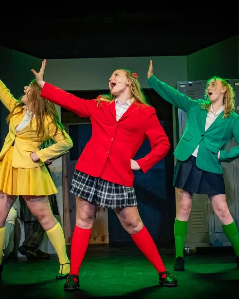 Three young women performing on stage with expressive poses, wearing colorful school-like uniforms, with green stage lighting