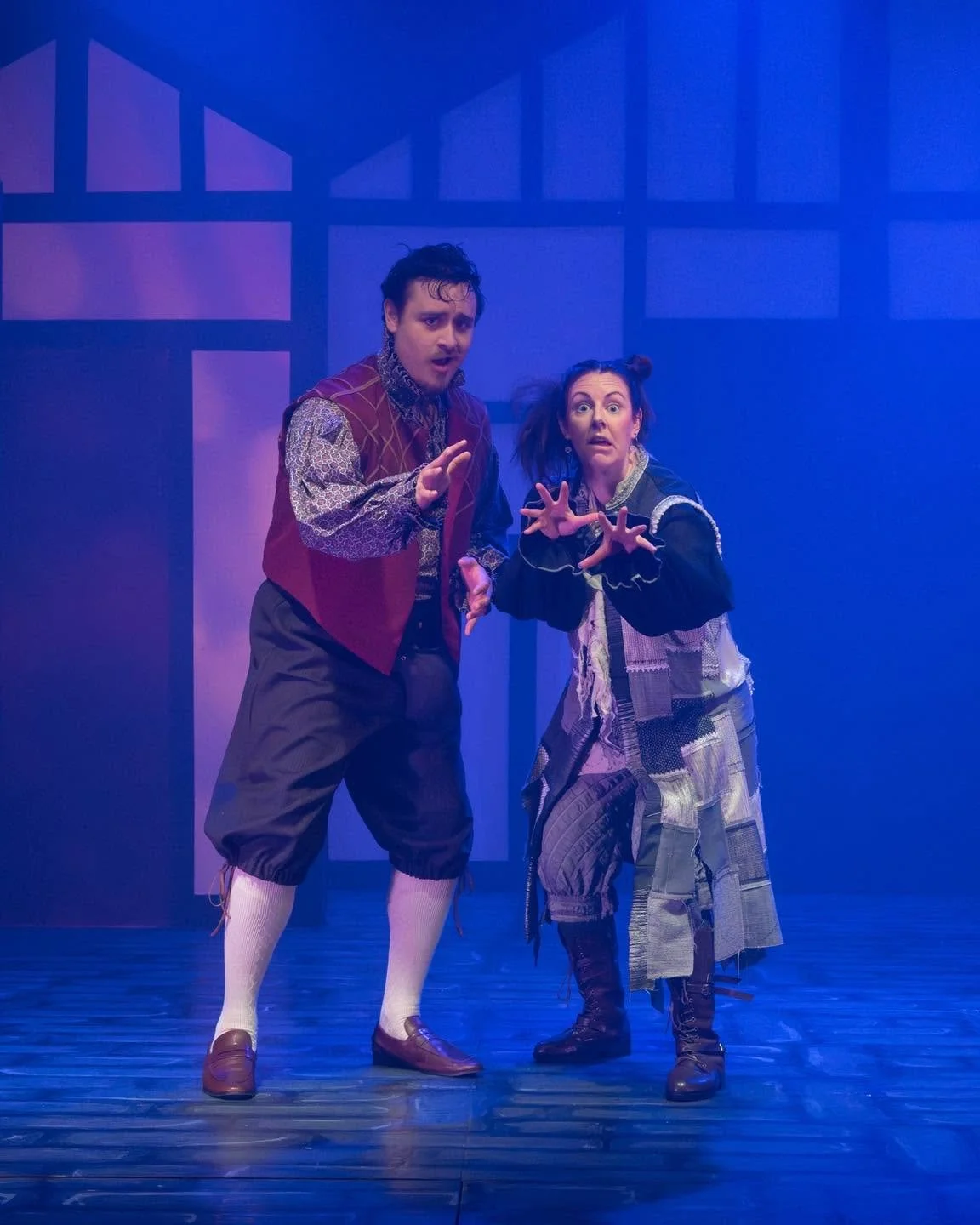 Two actors on stage with theatrical makeup and costumes, performing in a play with blue lighting and a minimalist set design.