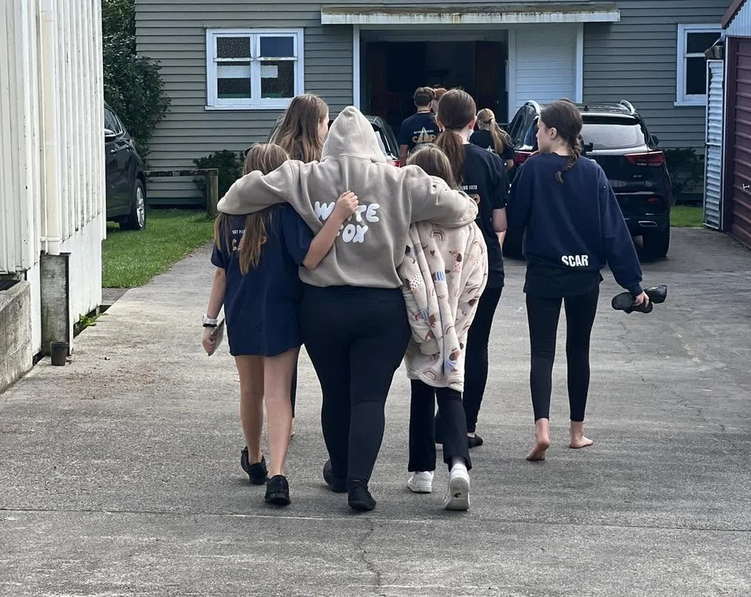 Group of children and a woman walking on a driveway towards a garage, all dressed casually in dark clothing, with some children holding objects, in front of a house with cars parked outside.