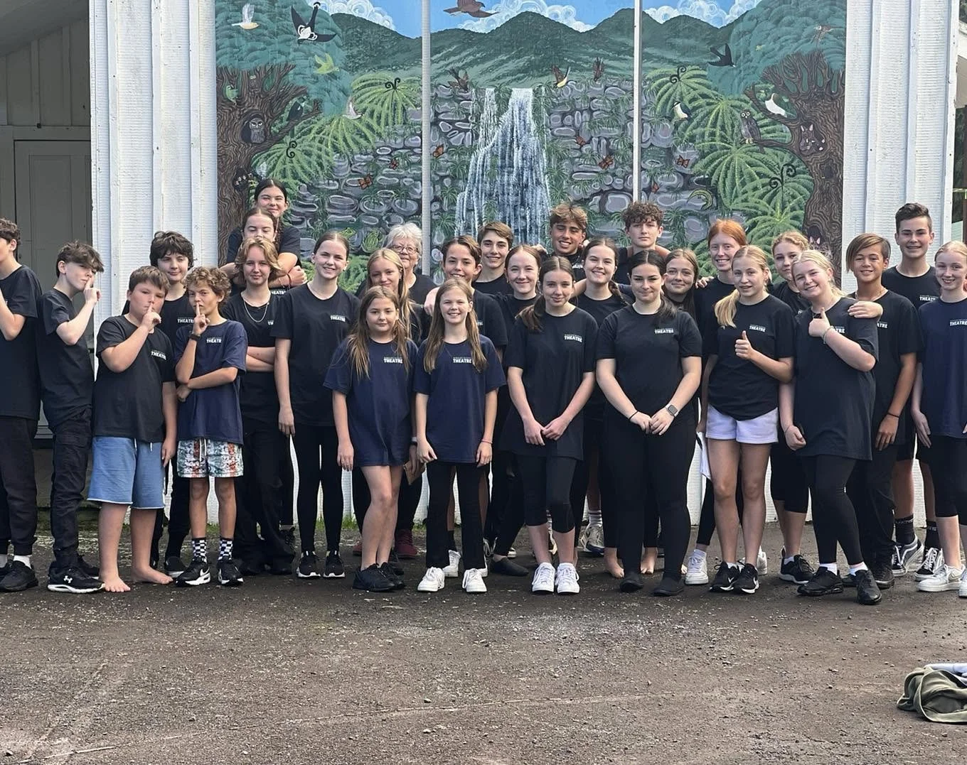 Group of children and teenagers standing outside in front of a colorful jungle-themed wall mural, some making funny faces and others smiling.