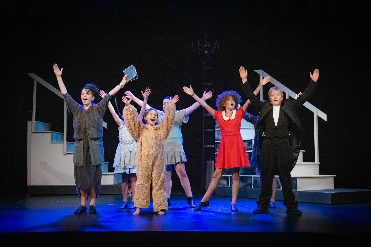 Children performing on stage with arms raised, dressed in costumes, with theatrical set pieces and stairs in the background.
