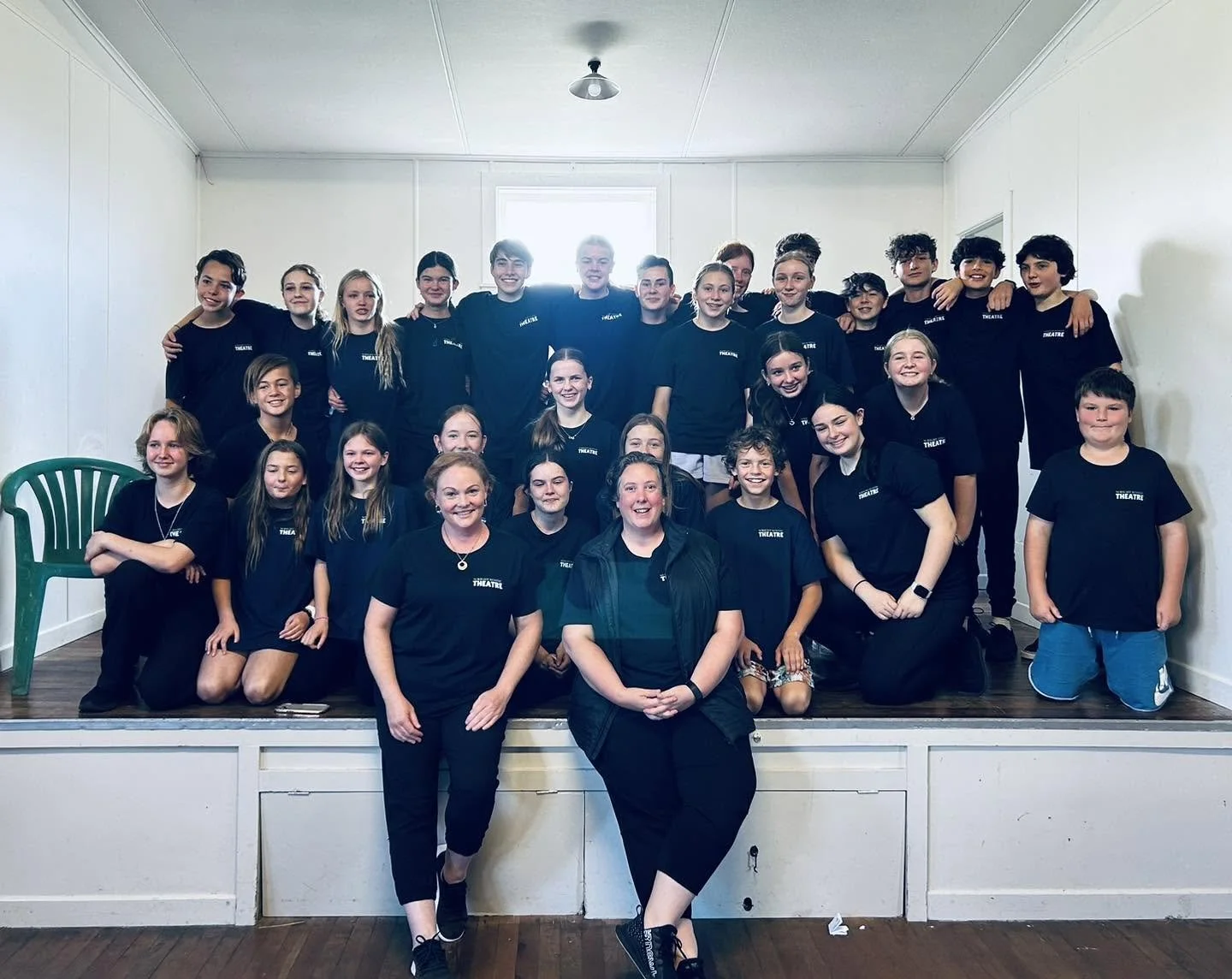 Group of children and two adults posing on a small stage in a room with white walls and natural light from a window behind them. They are all wearing black t-shirts with white text that says 'THEATRE'.