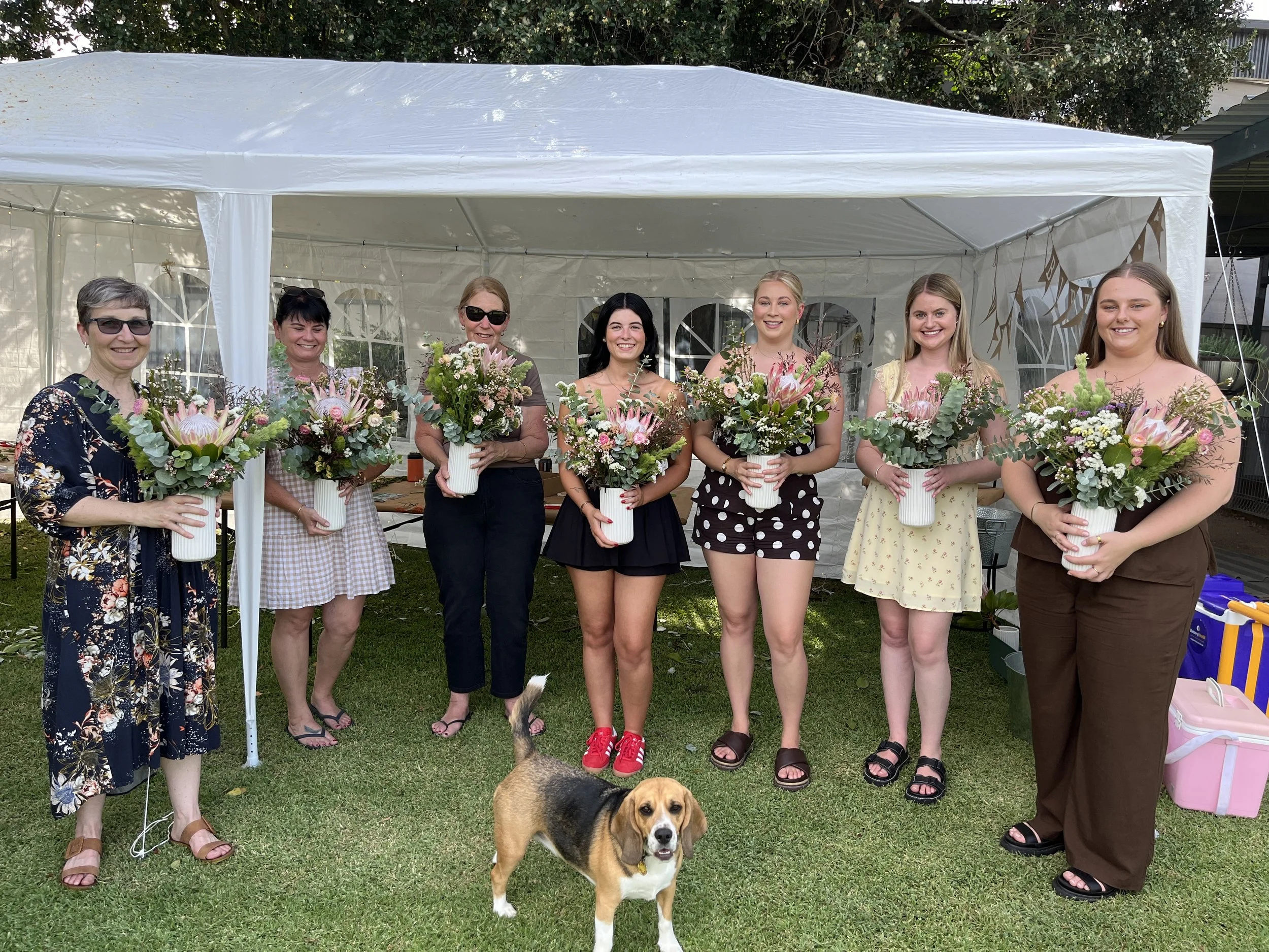 Seven women standing in a row outside under a white tent, each holding a bouquet of flowers, with a beagle dog in front. The women are smiling and dressed casually, with trees and party supplies visible in the background.