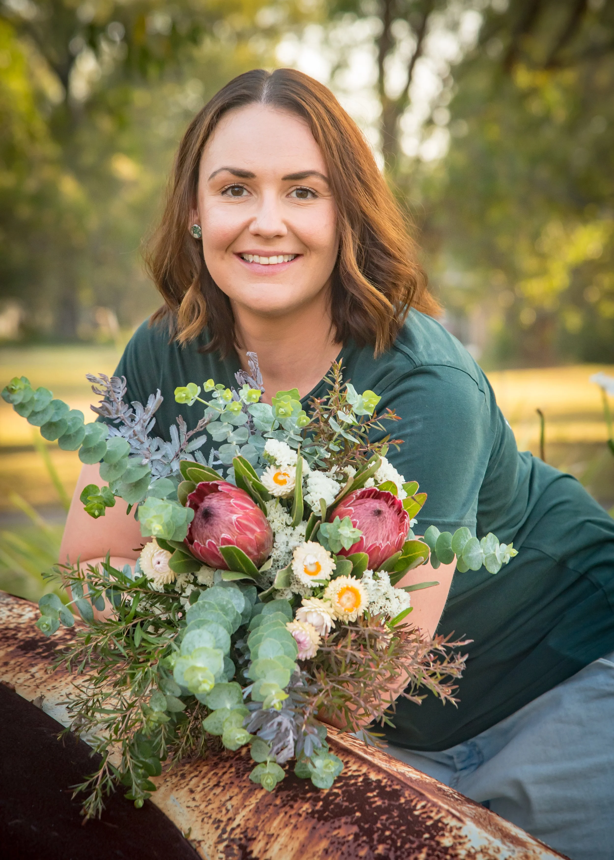 A woman with shoulder-length brown hair in a teal dress holding a bouquet of Native Australian and South African flowers outdoors, smiling at the camera with trees and sunlight in the background.