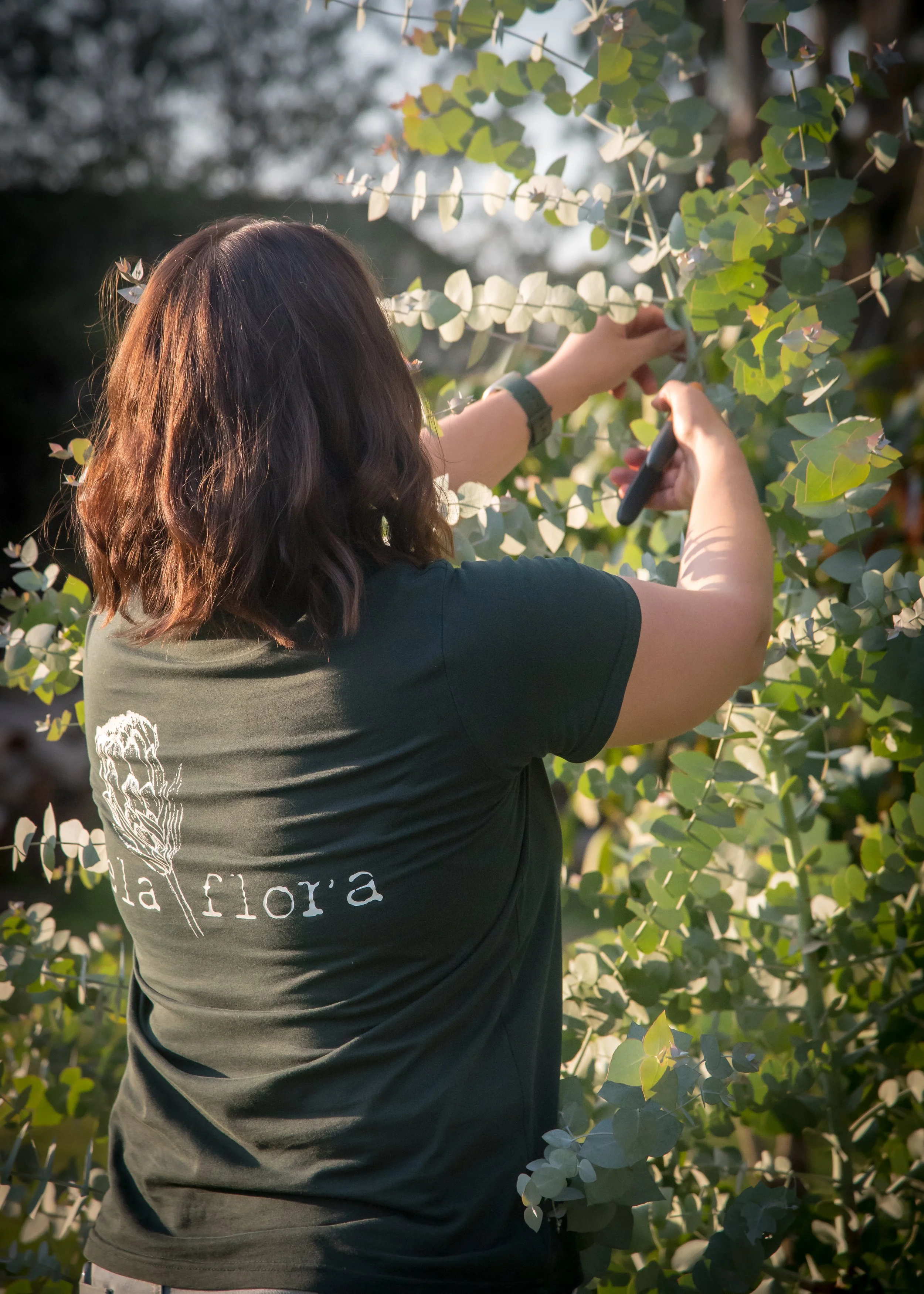 A woman with wavy brown hair, wearing a dark green t-shirt with a floral design and the words "Ela Flora," trimming a leafy green plant outdoors.