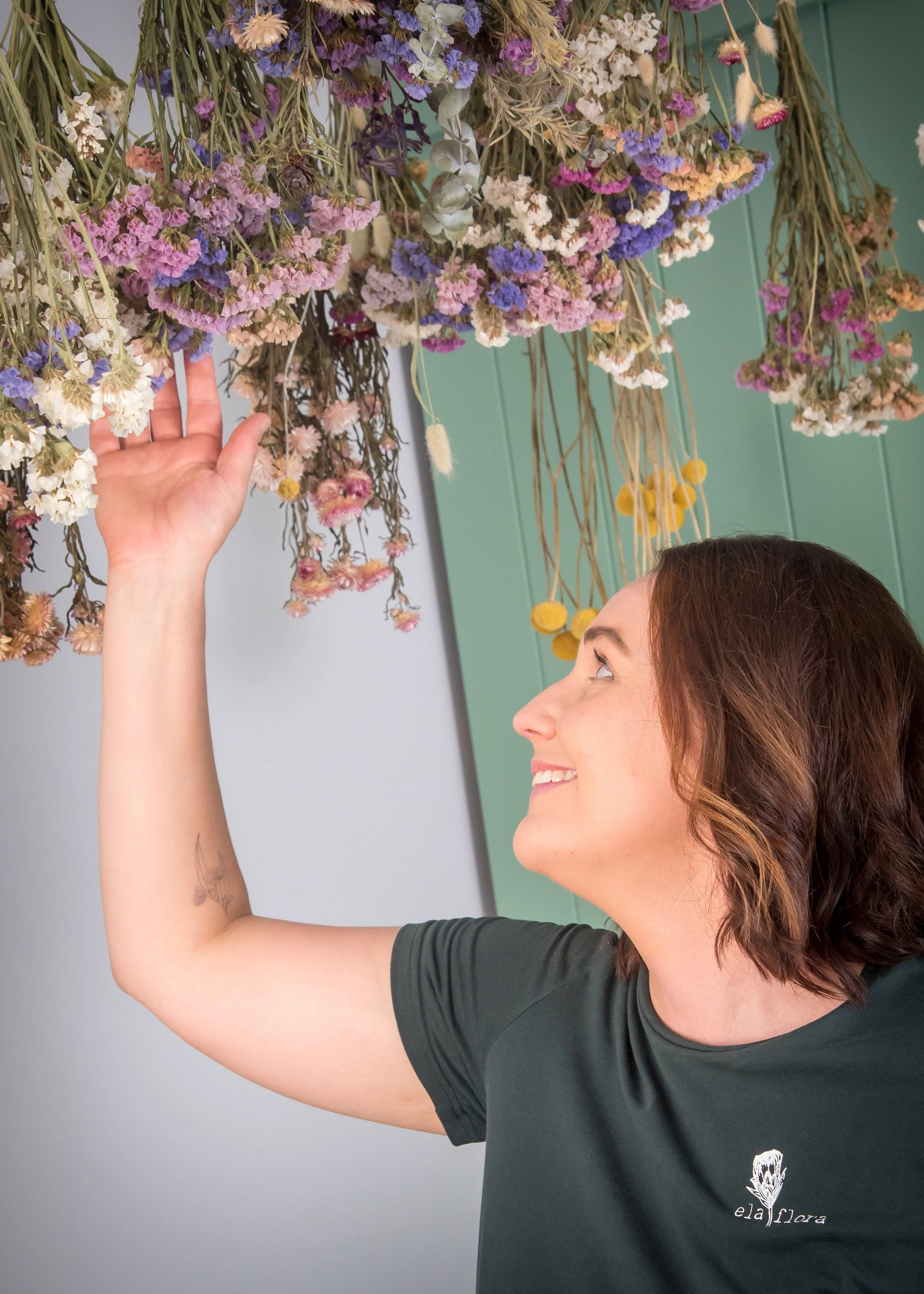 A woman with shoulder-length brown hair smiling and looking at a hanging bouquet of dried flowers in purple, pink, white, and yellow. She is wearing a dark green shirt with a small white logo on the chest and has a small tattoo on her left forearm.