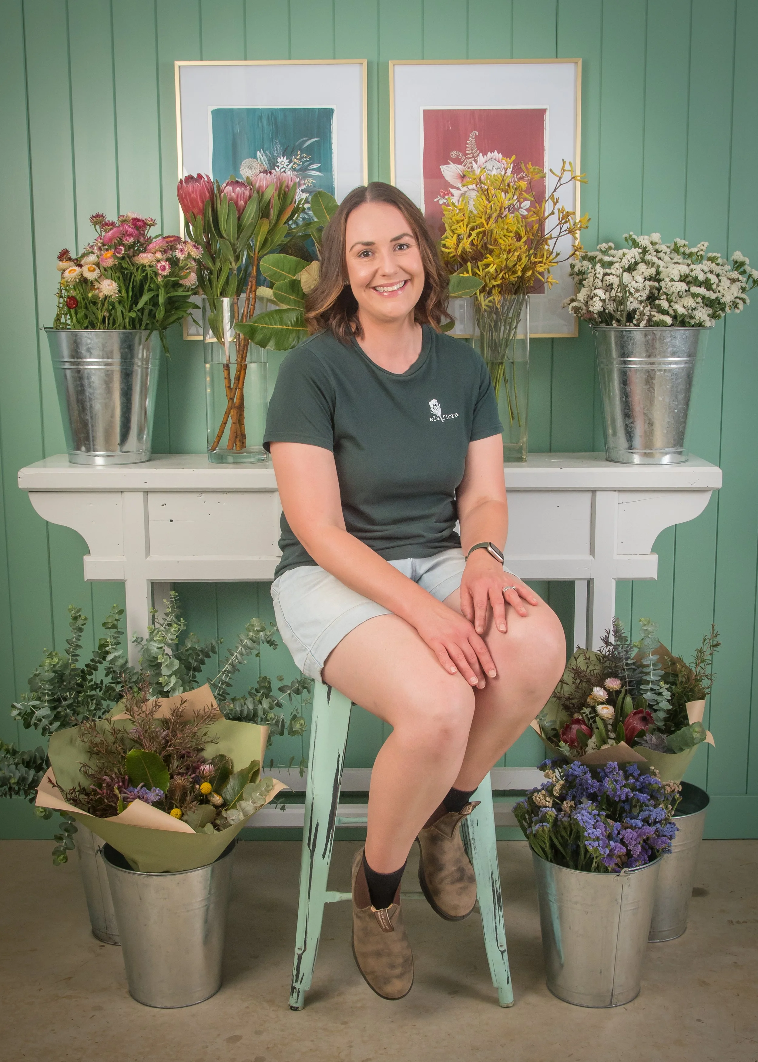 A woman sitting on a mint green stool in front of a white console table with five flower arrangements in metal buckets and vases, with colorful framed artwork hanging on a mint green wall behind her.