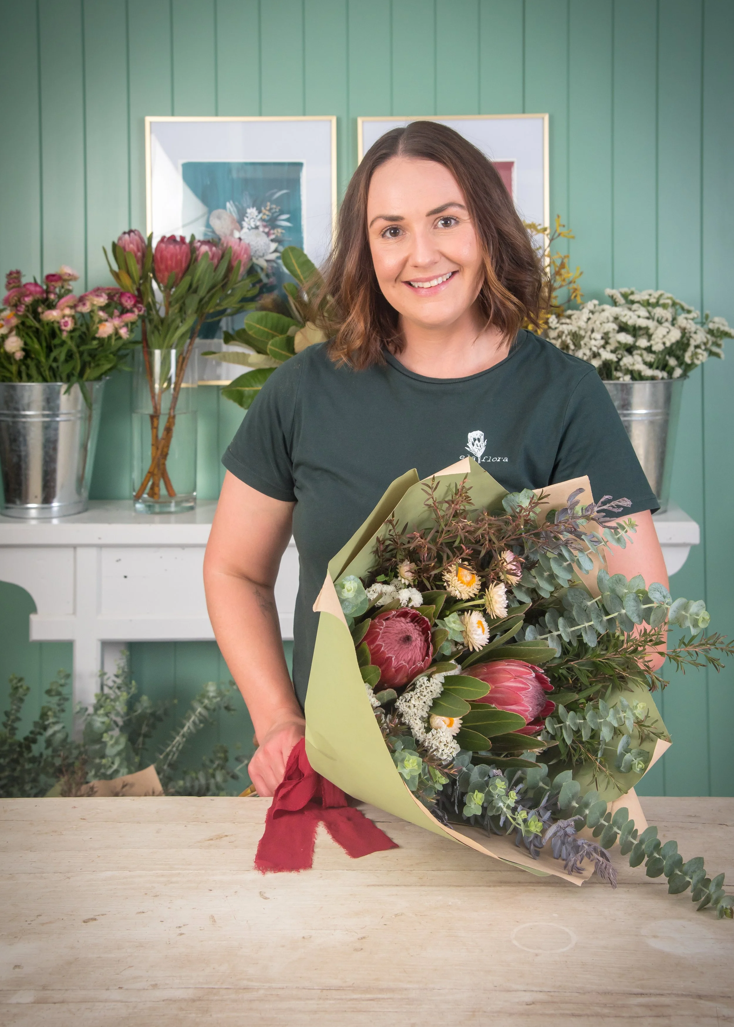 A woman in a dark green T-shirt smiling and holding a bouquet of flowers in a floral shop with assorted flowers and green wall decor in the background.
