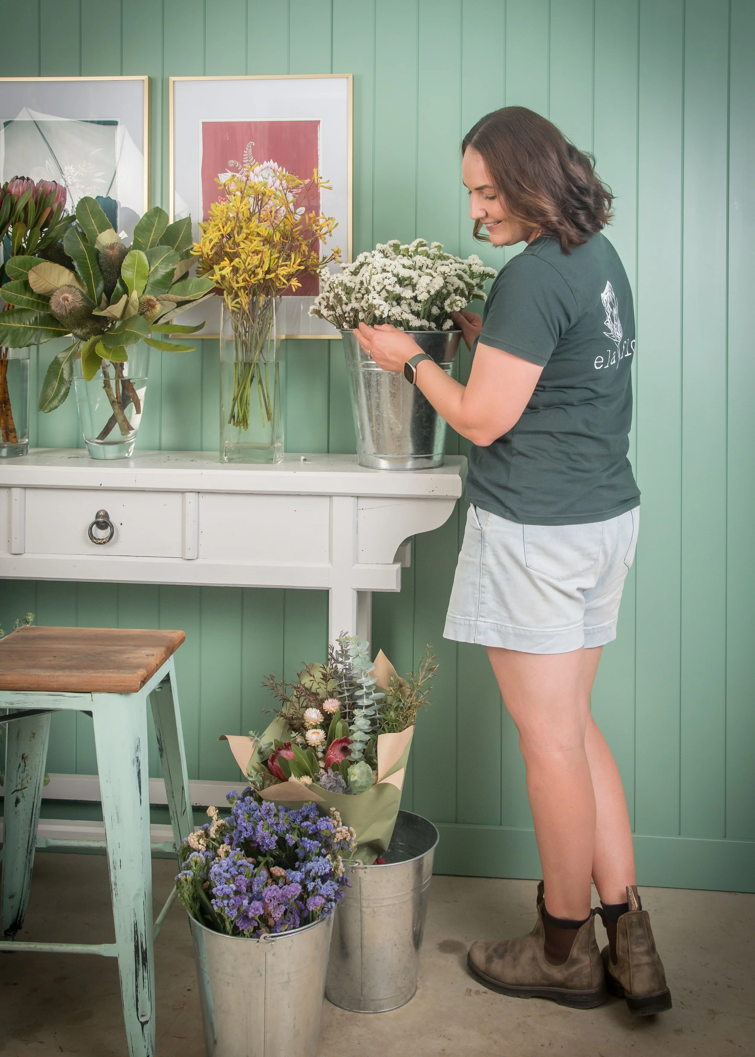 A florist in gray shorts and brown boots arranging white flowers in a silver bucket inside a floral shop. There are potted purple and pink flowers on the floor and various flower arrangements on a white table against a green paneled wall.