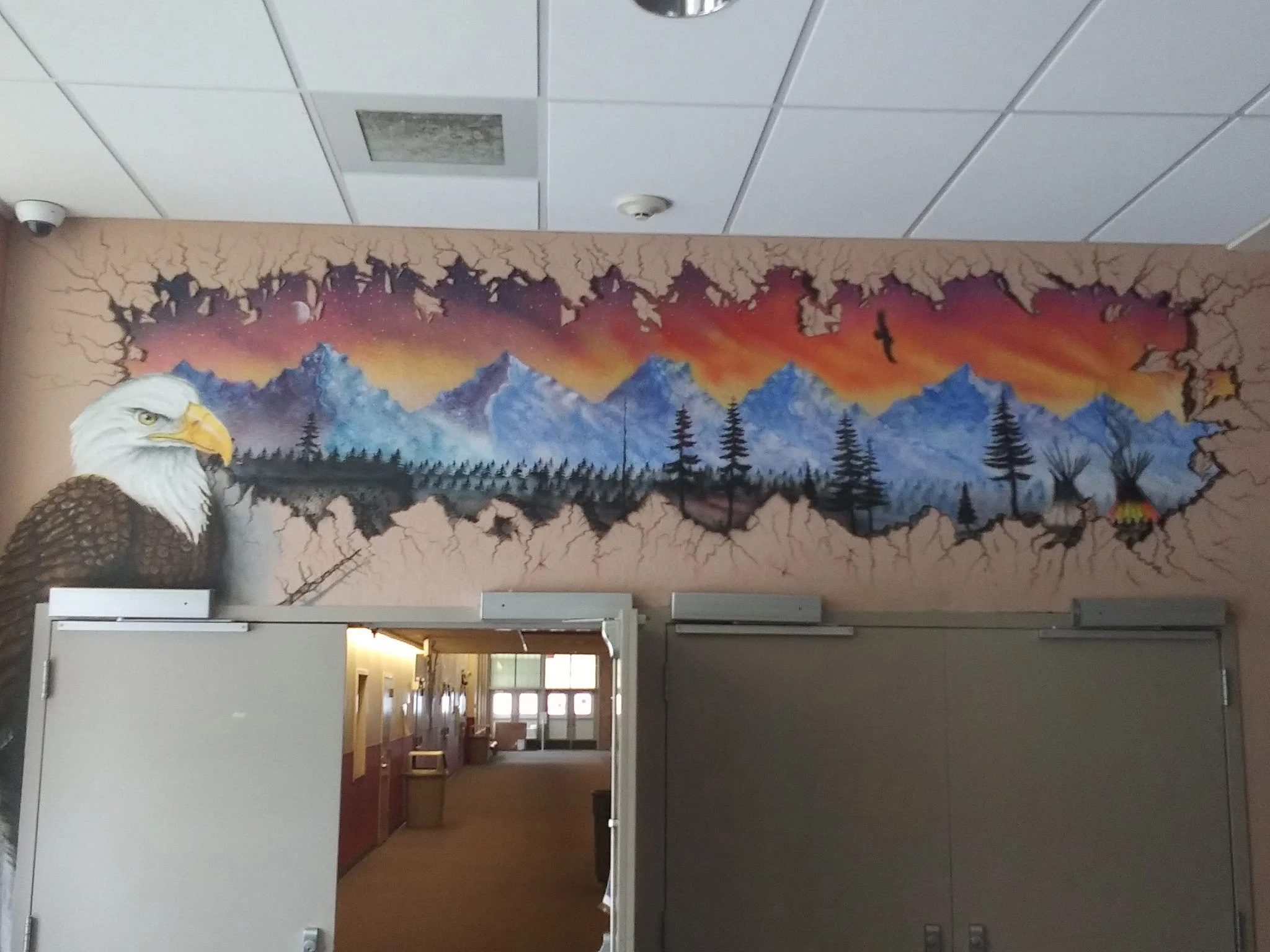 A mural painted above the door to a restaurant. The mural depicts a mountain range skirted by trees and bushes and a sunset in the background.