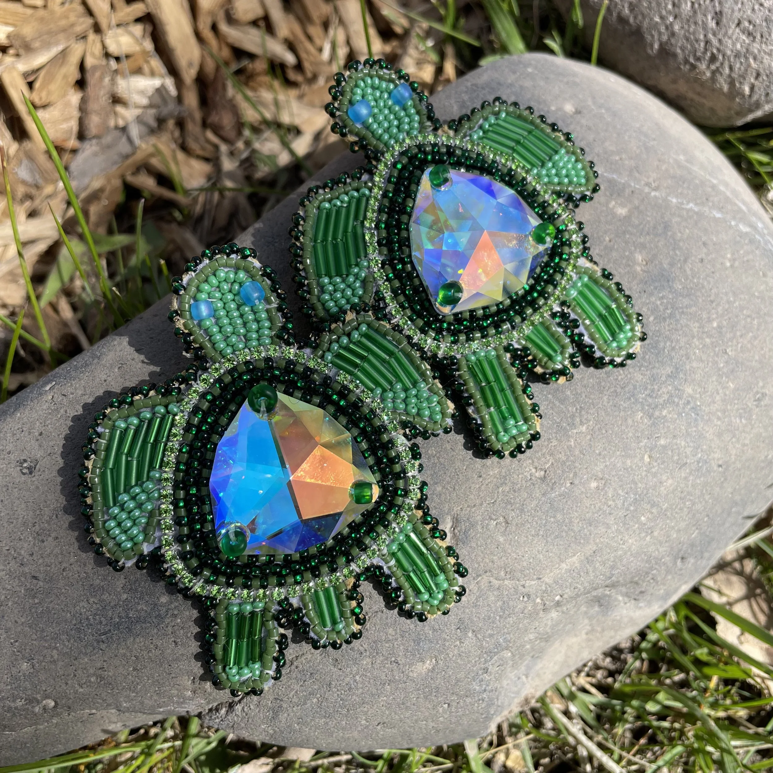 Pair of green beaded earrings with large iridescent stones in the center, resting on a gray stone surface outdoors.