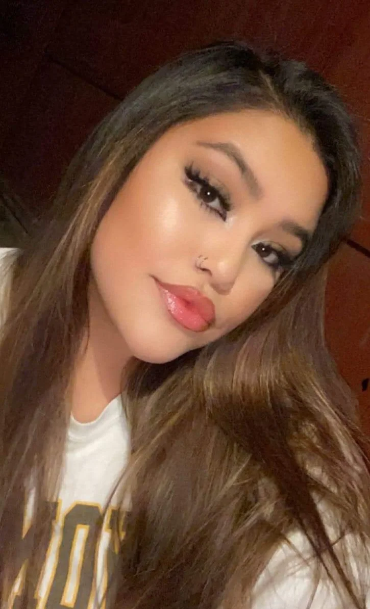 Close-up of a woman with long brown hair, makeup, hoop nose ring, sticking out her tongue, and wearing a white top.