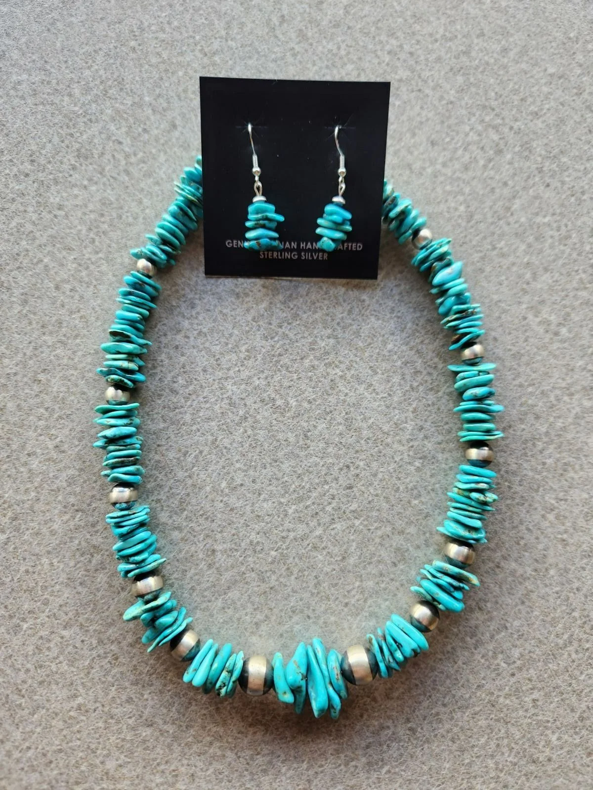 A turquoise and silver jewelry set, including a necklace and matching earrings, made of turquoise chips and silver beads, displayed on a plain surface.