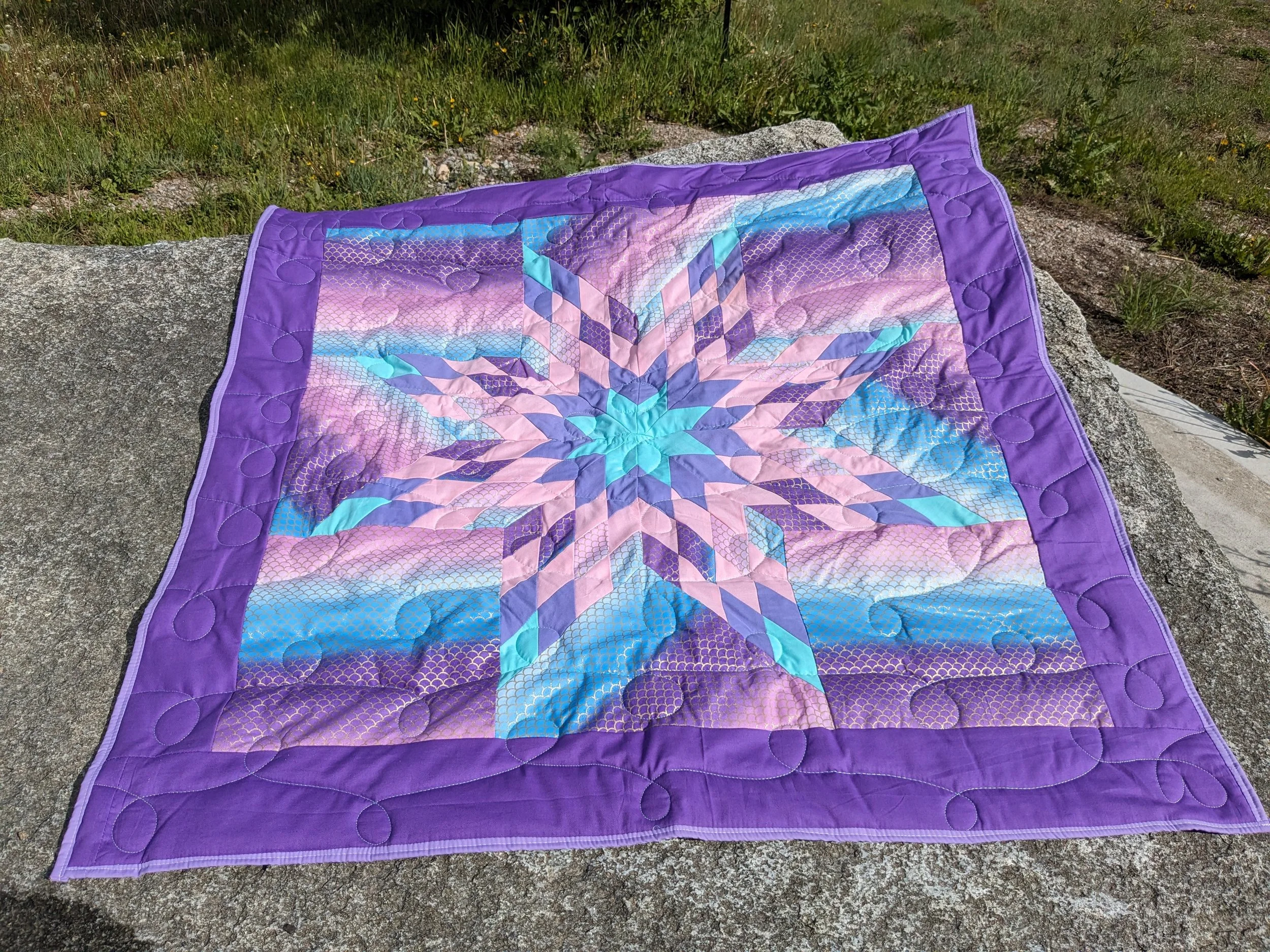 A colorful quilt with a star-like pattern in shades of purple, pink, and blue, laid on a large rock outdoors.