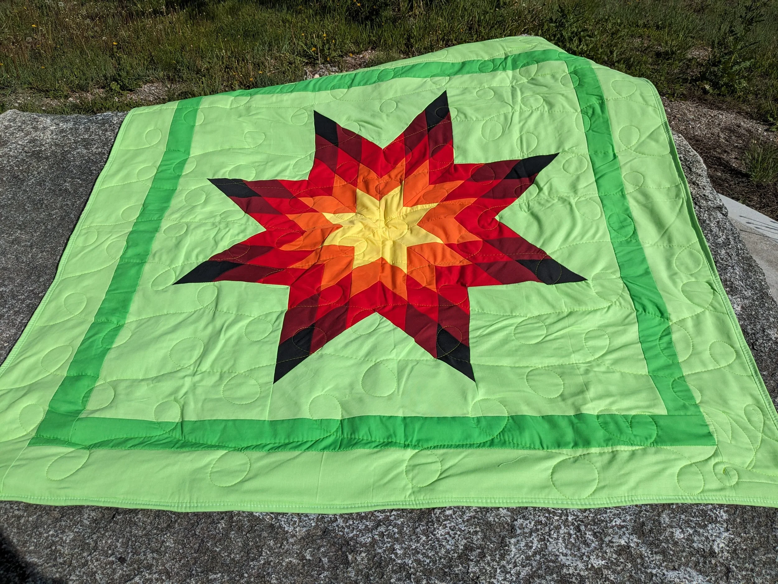 A brightly colored quilt with a star pattern placed on a large rock outside.