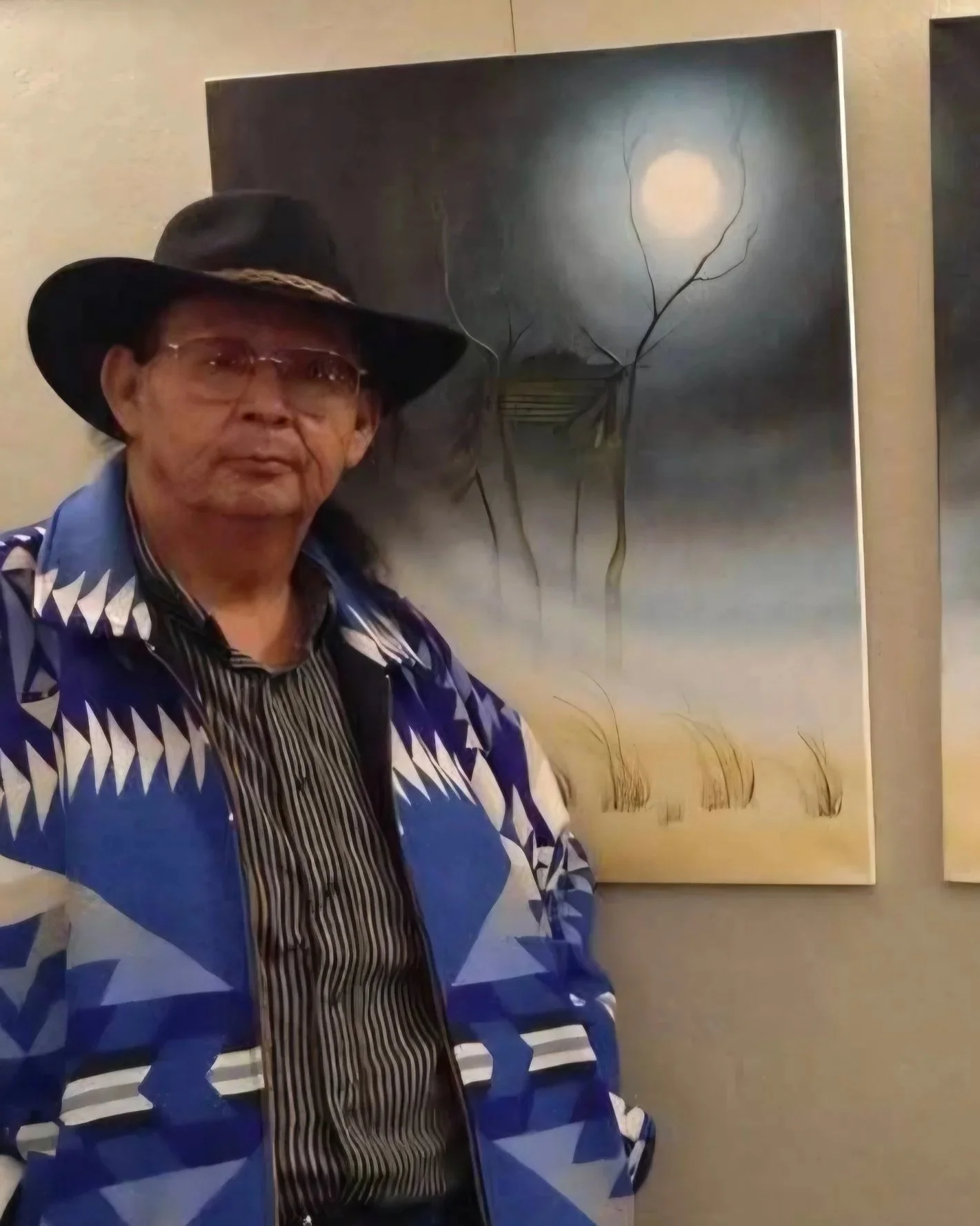 An older man wearing a black hat, glasses, a striped shirt, and a blue patterned jacket stands in front of a painting depicting a moonlit scene with a tree and tall grass.