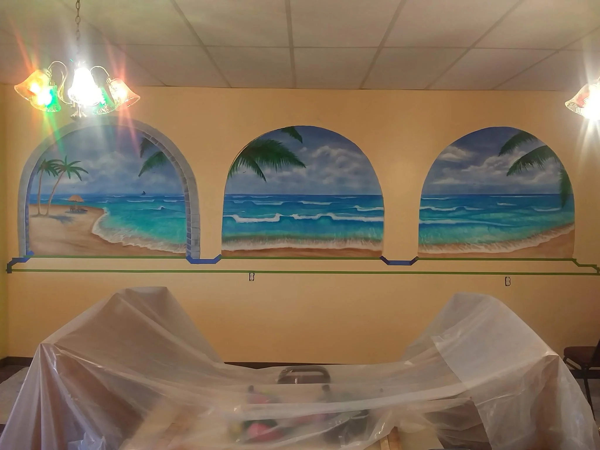A painted mural in three arched inlays in a restaurant. The 3 paintings portray 3 views of a beach with the ocean lapping onto the sand. There are palm trees in the farthest left arch.