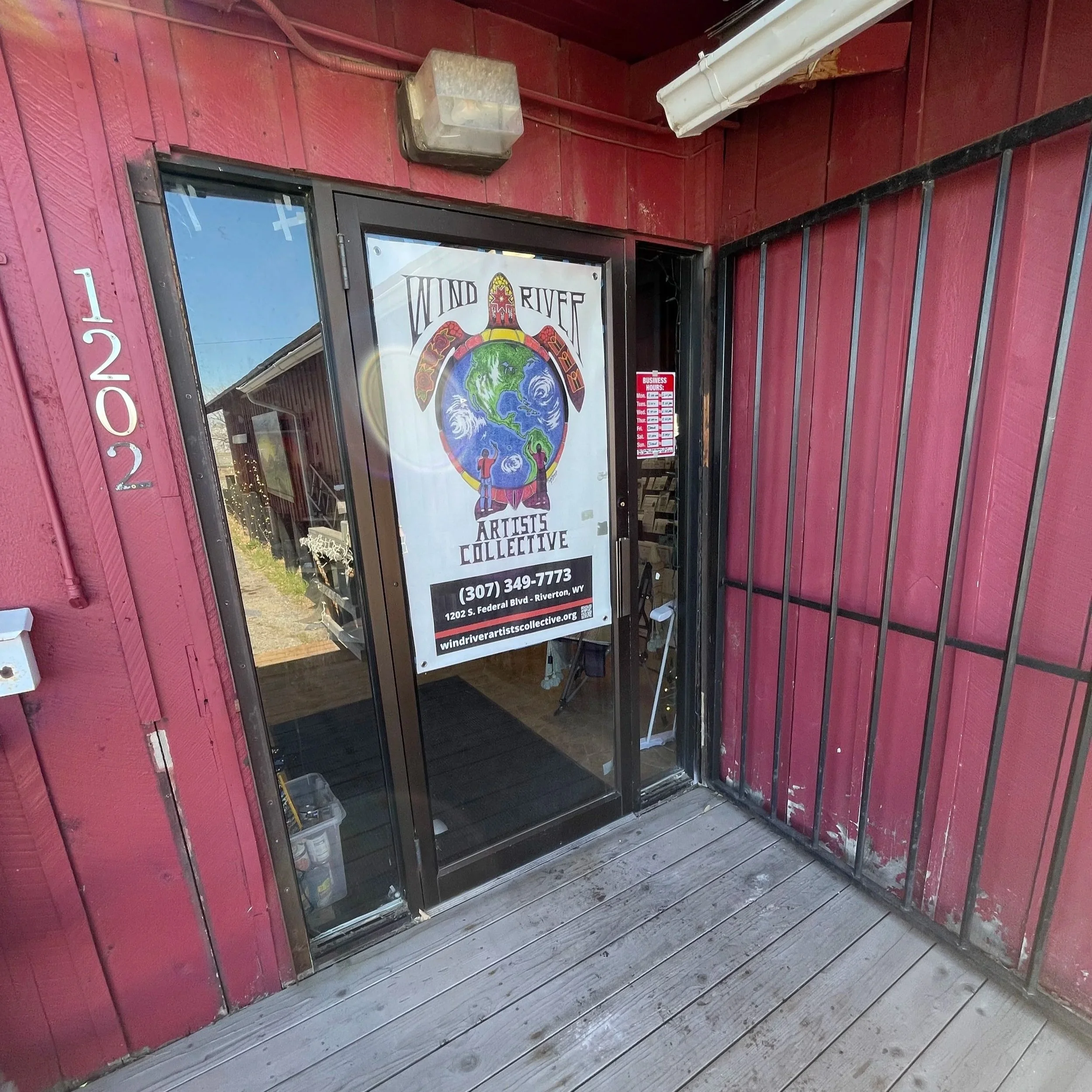 Entrance of Wind River Artists Collective with a glass door displaying a colorful poster, and on the left, the building's red wood siding with the number 1202 vertically aligned.