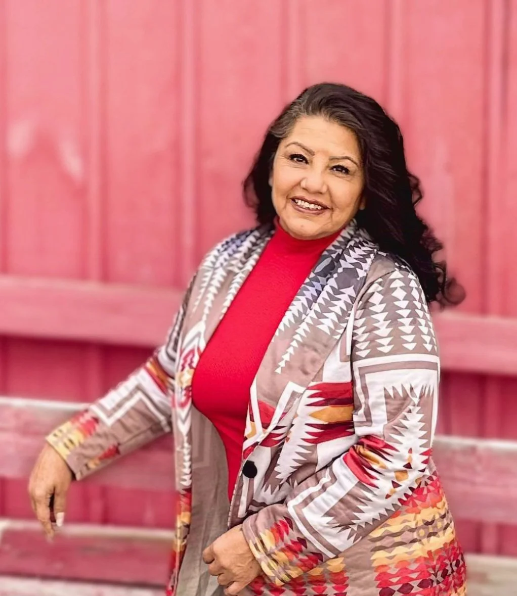 A smiling woman with dark hair, wearing a red top and a patterned jacket, stands in front of a pink wooden background.