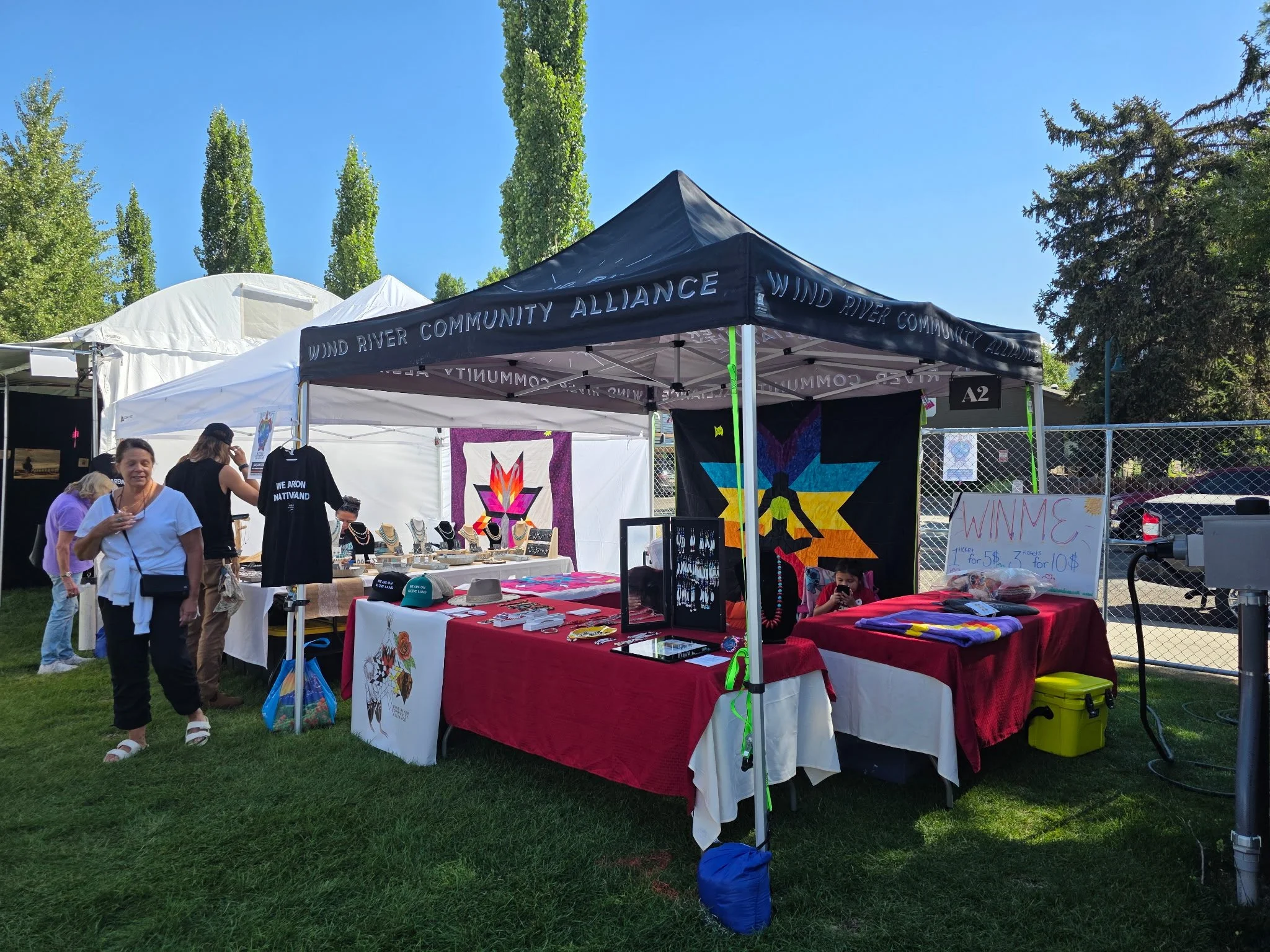 Outdoor art and craft booth at a fair or market, with display tables covered with various handmade items, jewelry, and t-shirts. There are several people browsing and working at the booth, with colorful banners and art pieces on the tent and tables. Trees and a chain-link fence are visible in the background.