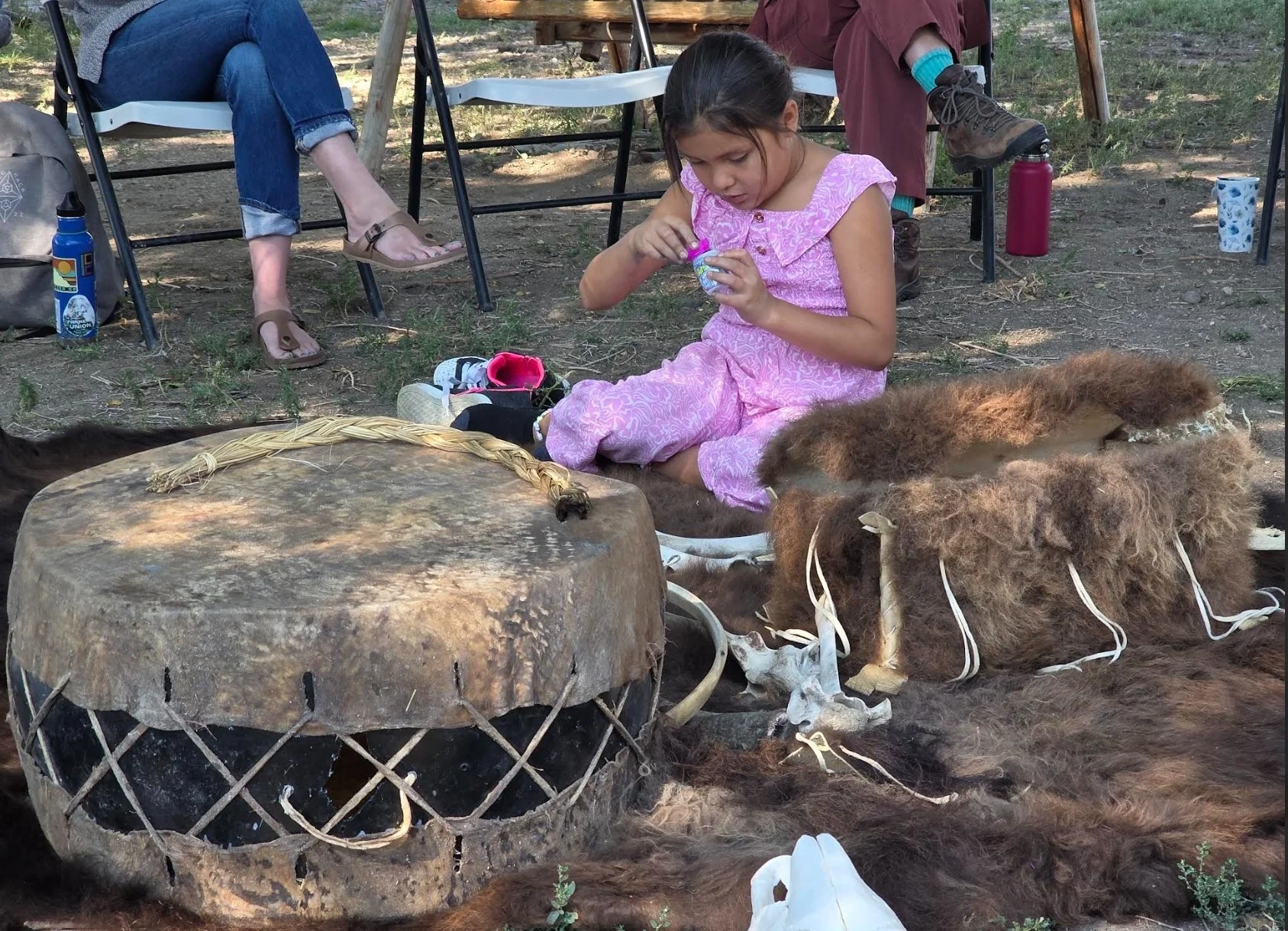 A young girl in a pink dress sits on the ground, opening a small plastic container, surrounded by brown furry animal hides and skulls, with an outdoor fire pit in the foreground and two adult people sitting on chairs nearby.