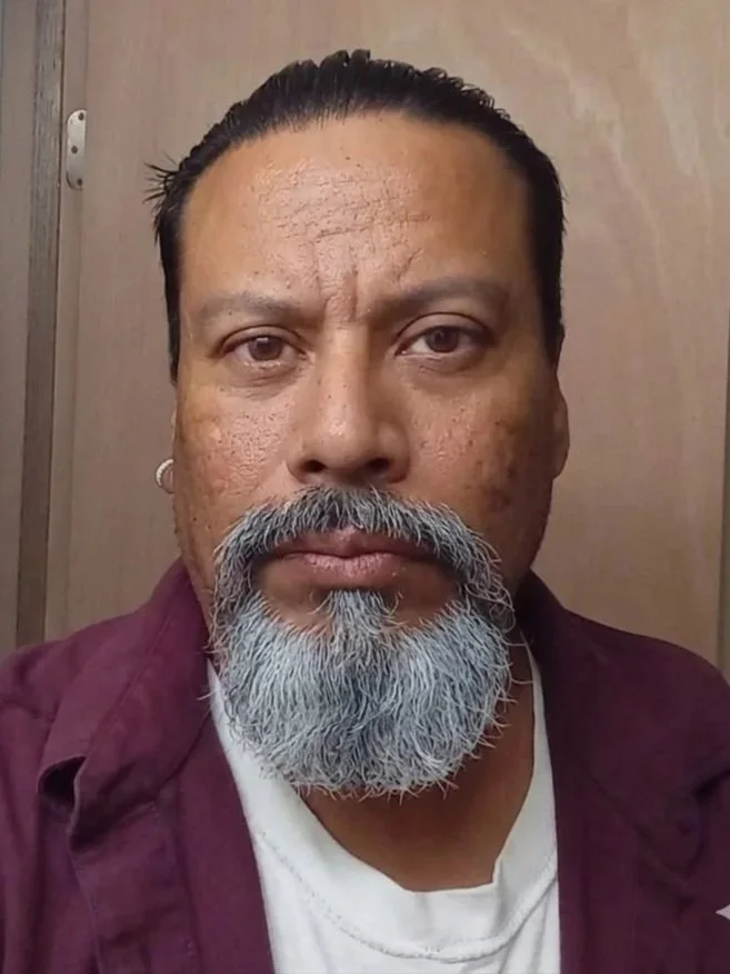 A close up selfie of a man wearing a maroon button up and a white undershirt. The man has black hair pulled back, a salt and pepper beard, and a piercing in his right earlobe