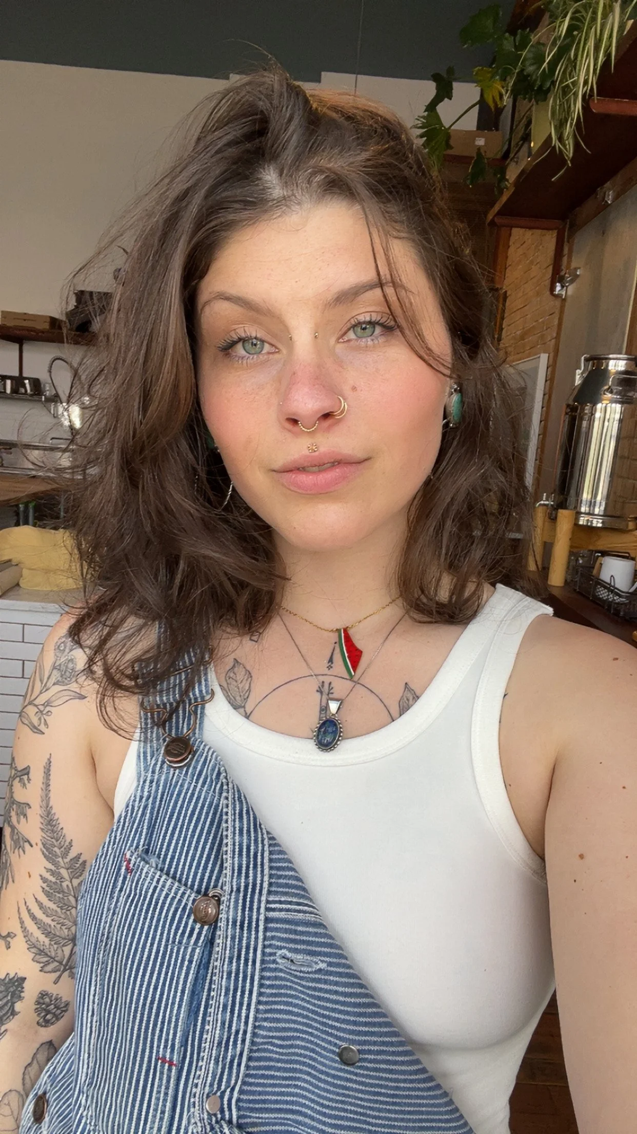 A young woman with light skin, wavy brown hair, and blue eyes taking a selfie indoors. She has multiple facial piercings and tattoos on her arm, and is wearing a white tank top, layered necklaces, and a denim overall strap on her shoulder.