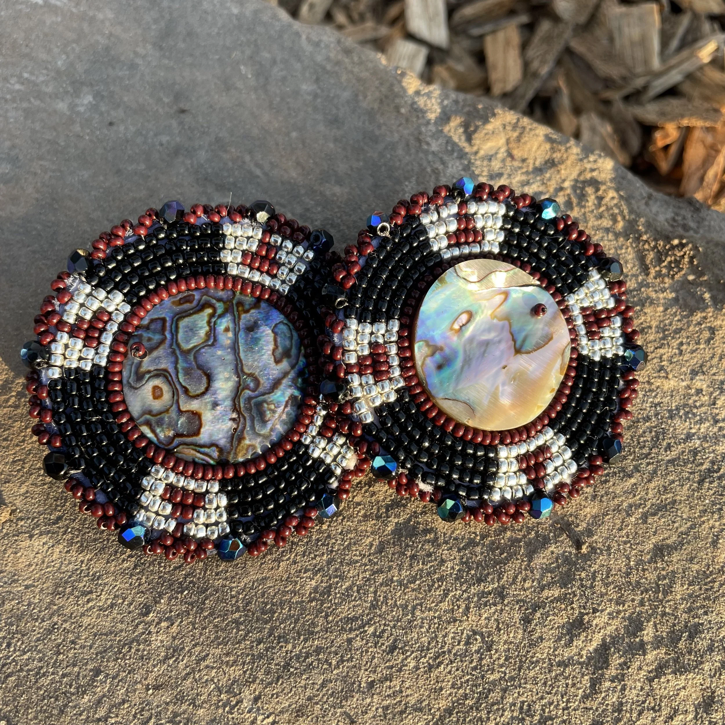 Two intricately beaded jewelry pieces featuring large iridescent shell or mother-of-pearl centers, with black, red, silver, and blue beads forming circular patterns.