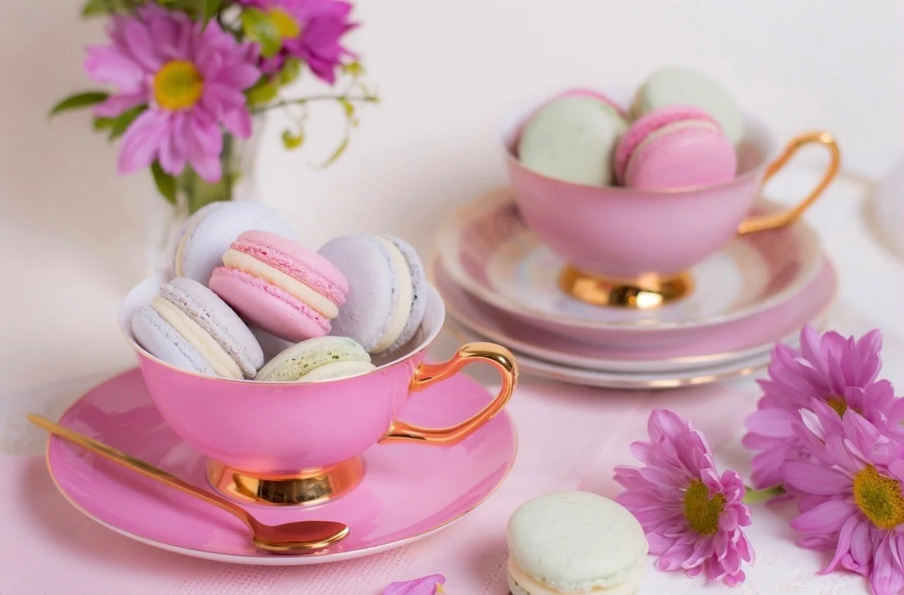 Two pink tea cups filled with colorful macarons, placed on matching pink plates with gold accents. Surrounding flowers, including pink daisies, and a pink spoon are also visible.