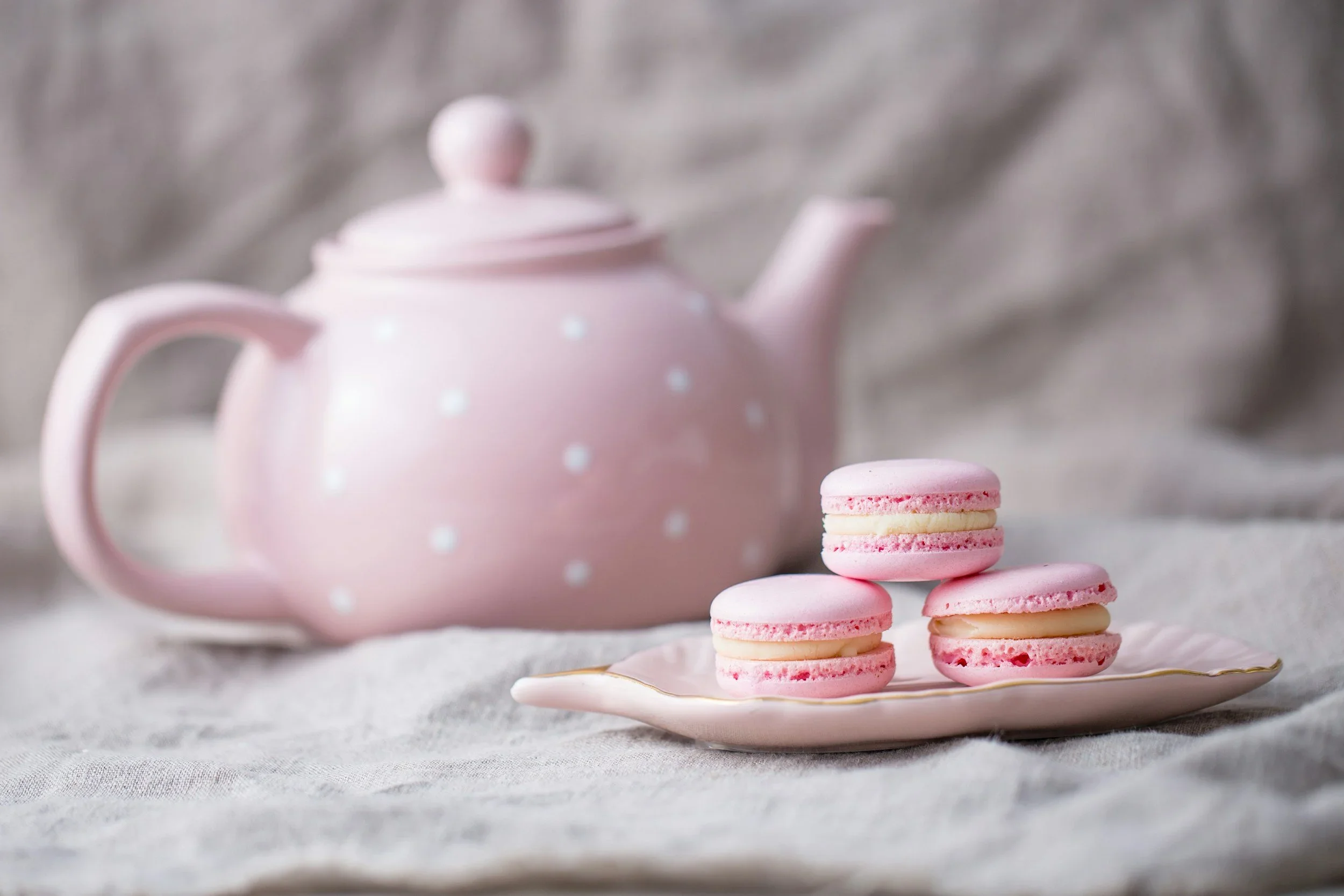 A pink teapot with white polka dots and a matching lid next to a small stack of pink and white macarons on a decorative plate, against a soft, textured background.