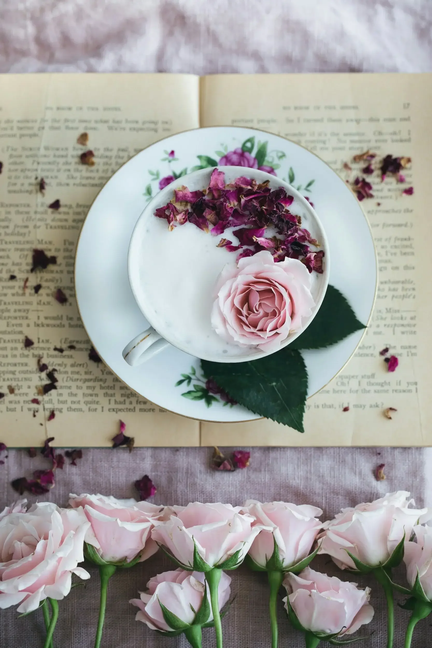 A teacup with pink roses and dried flower petals on a floral plate, placed on an open book, with pink roses at the bottom of the image and scattered dried petals around.