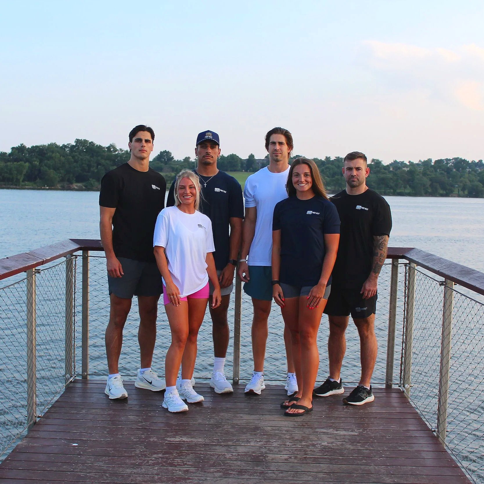 Group of six young adults standing on a wooden dock by a body of water with a treeline in the background, wearing high-quality athletic clothing.
