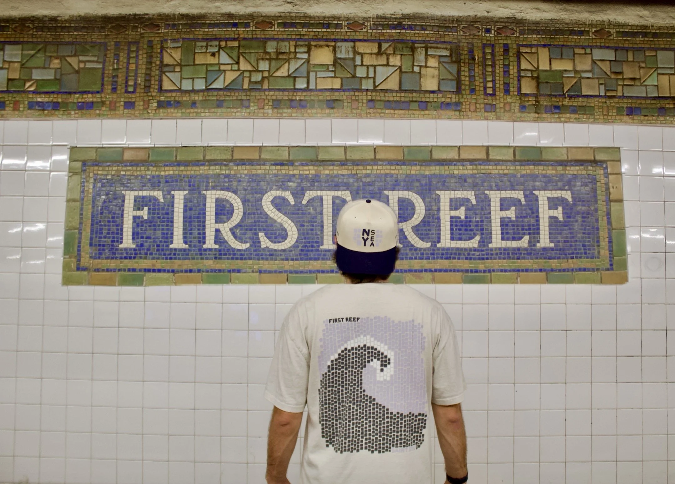 Person standing in front of a New York City Subway mosaic wall with the words 'FIRST REEF' in large blue letters.